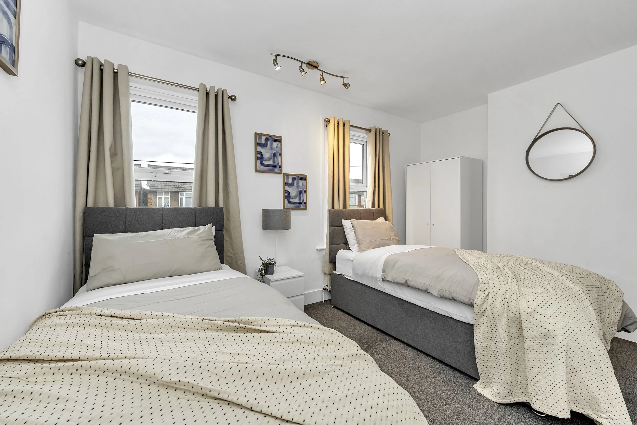 A bedroom with two twin beds, neutral-colored bedding, and beige curtains. There are two windows, one behind each bed, with a small white bedside table and a lamp between them. Decor includes three framed blue and gold abstract art pieces on the wall and a modern mirror on the right wall.