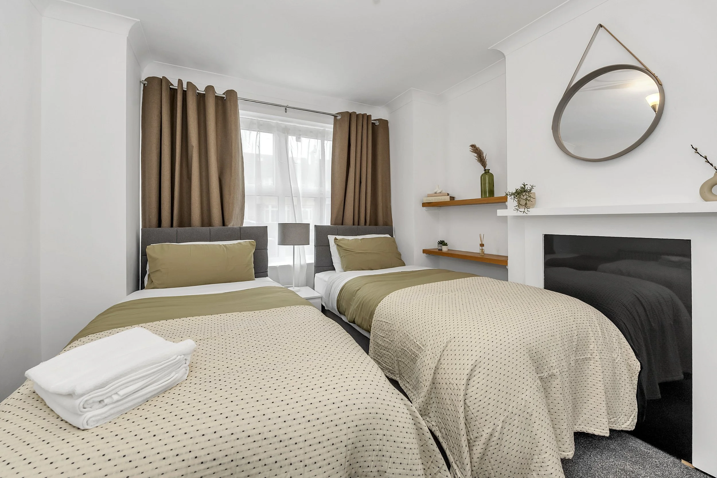 A cozy bedroom with three beds, each with beige and gray bedding, a window with white curtains and brown drapes, a white wall with a round mirror, a fireplace, and two floating wooden shelves with decorative items.