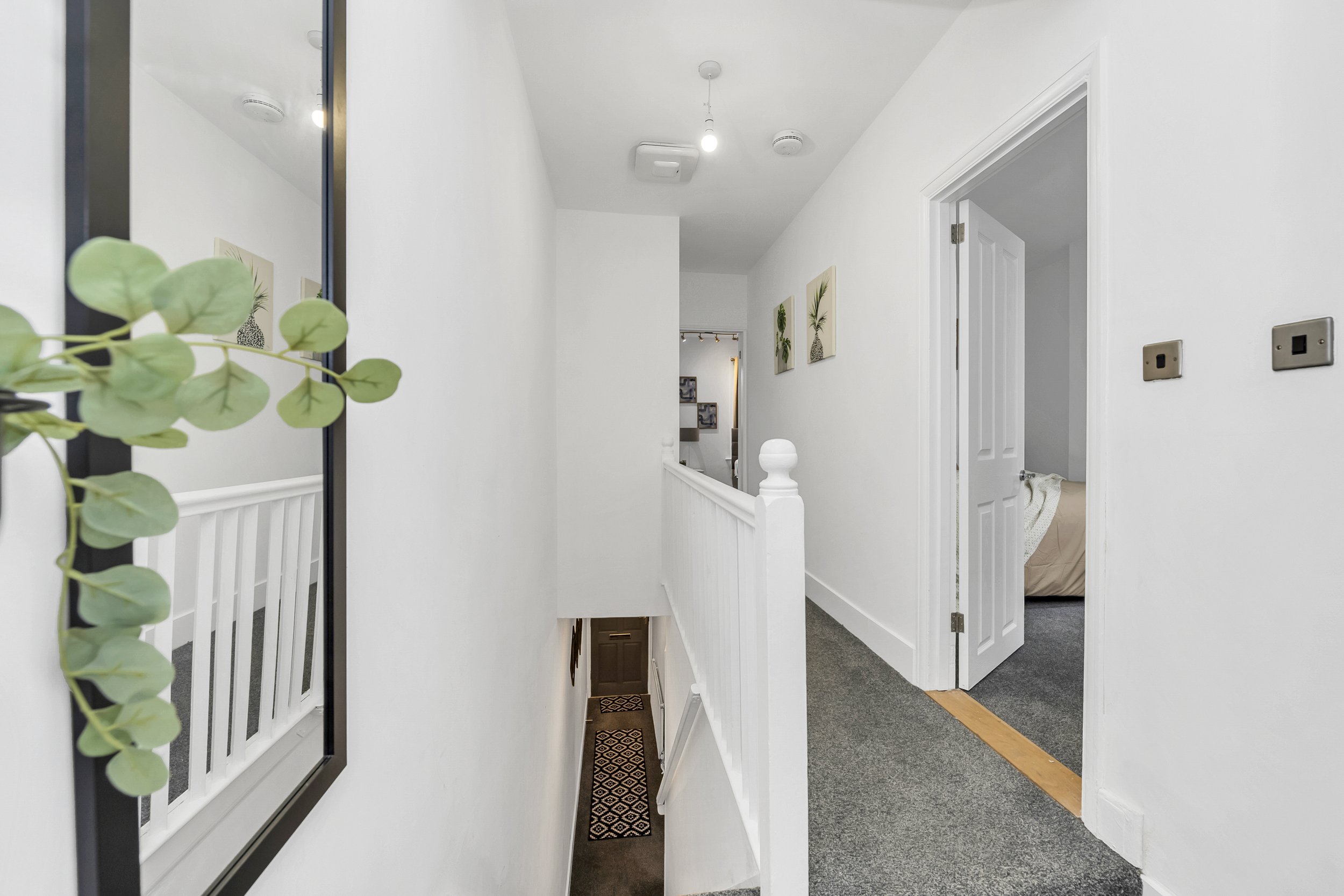 Interior of a white residential hallway with a staircase leading downstairs, a framed picture on the wall, and an open door to a bedroom.