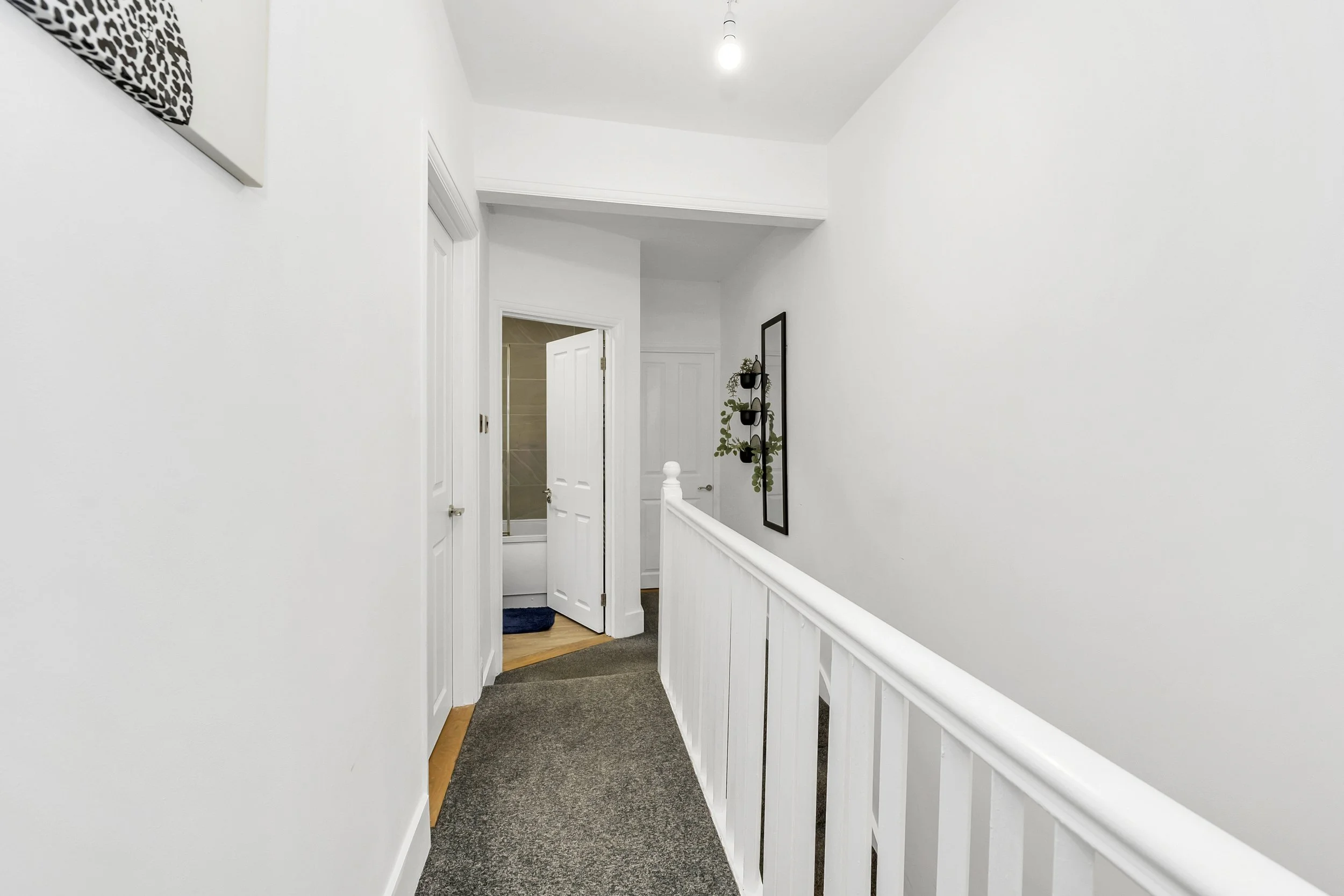 A white hallway with a staircase railing on the right, an open door to a bathroom with a bathtub, and a wall with a vertical black-framed mirror and some hanging plants.