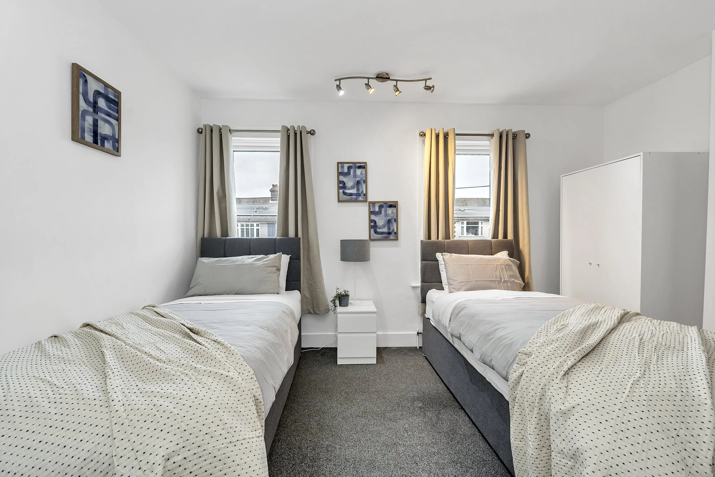 A bedroom with two twin beds with gray upholstered headboards, a white nightstand, a gray table lamp, a small potted plant, two windows with curtains, and framed abstract art on the walls.