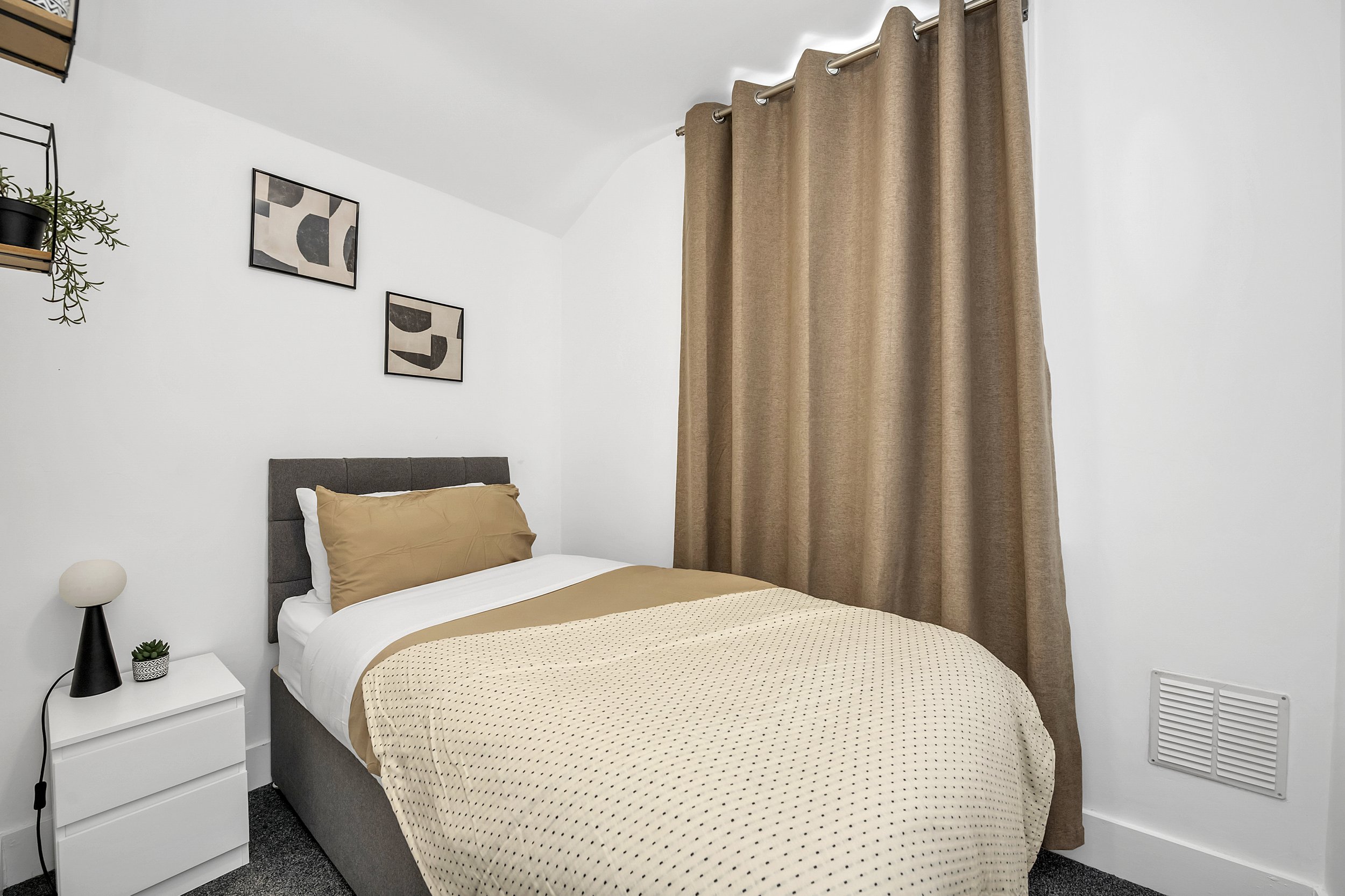 A neatly made bed with a grey upholstered headboard, beige and white bedding, and two beige pillows in a modern bedroom. There is a white nightstand with a small lamp and a potted plant. Beige curtains cover a window, and abstract art prints hang on the white wall above the bed.