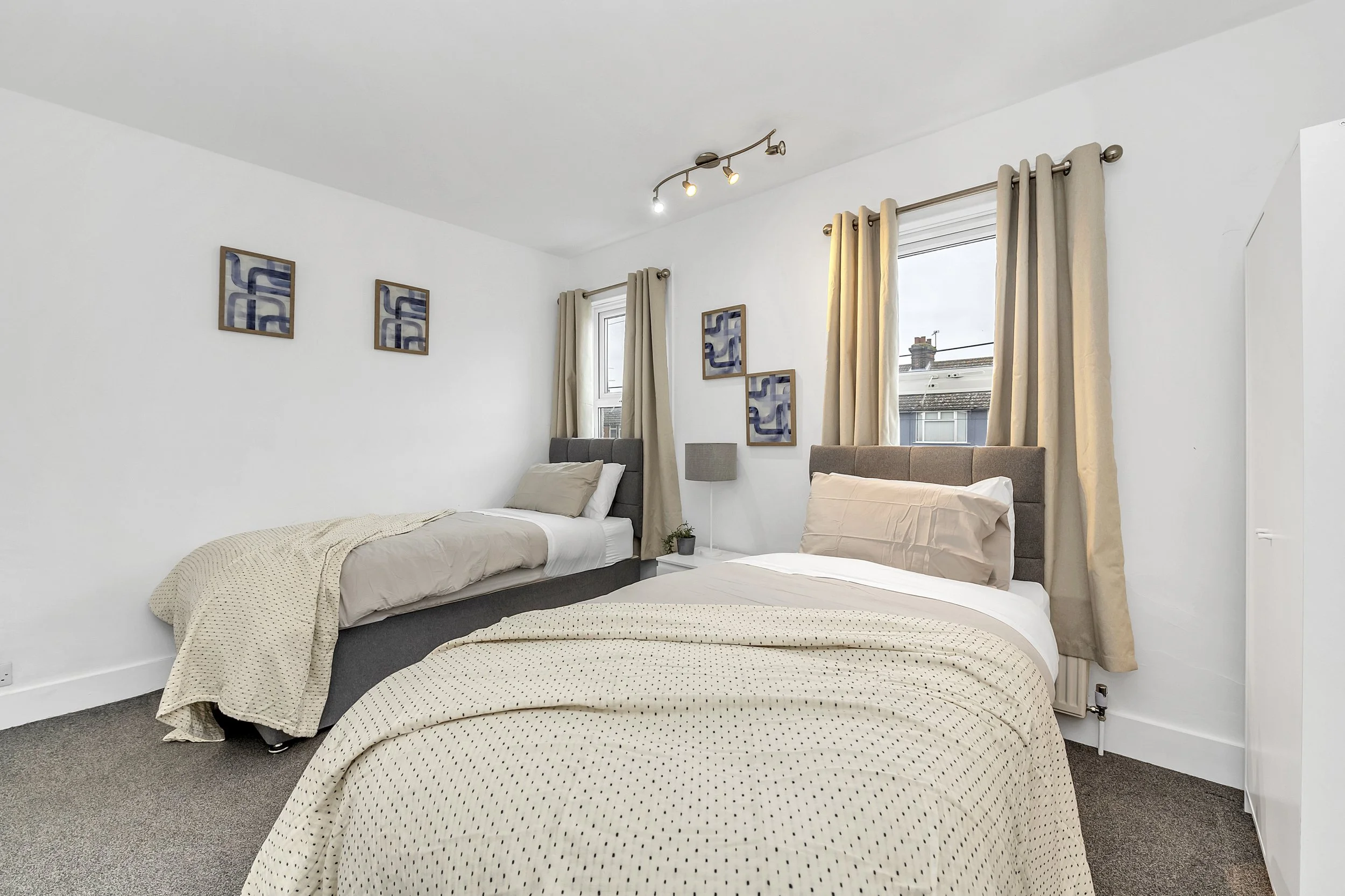 A bedroom with two single beds, beige and gray bedding, a small nightstand with a lamp, framed wall art, and windows with beige curtains.