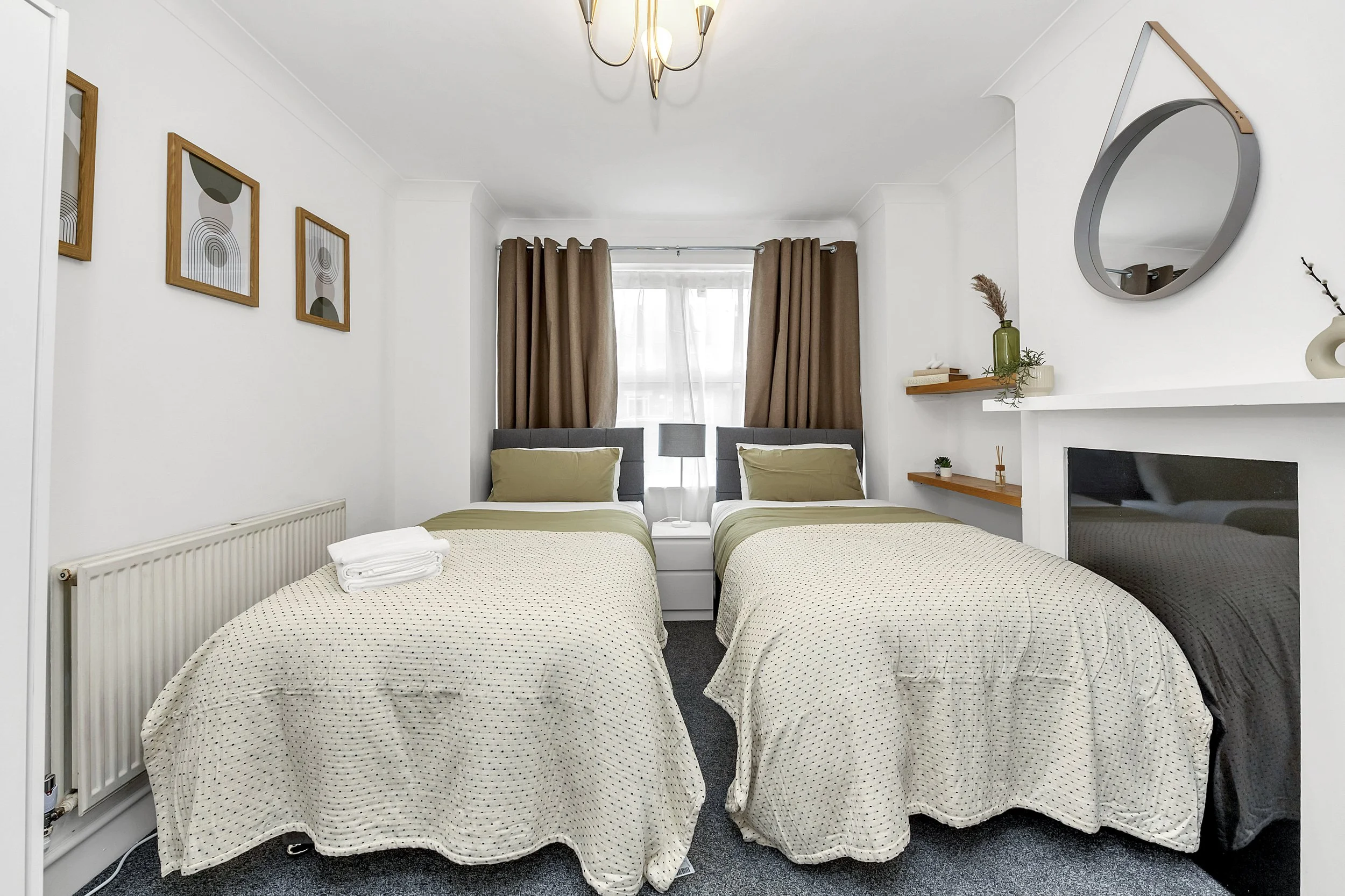 A bedroom with two single beds, each covered with beige and green bedding, placed side by side with a nightstand between them. The nightstand has a lamp. There are three framed abstract art pieces on the left wall. The window behind the beds has brown curtains. A small shelf on the right wall holds decorative items, including a vase with dried flowers. A mirror is mounted above the fireplace on the right, and there are small potted plants and decorative objects on the shelf.