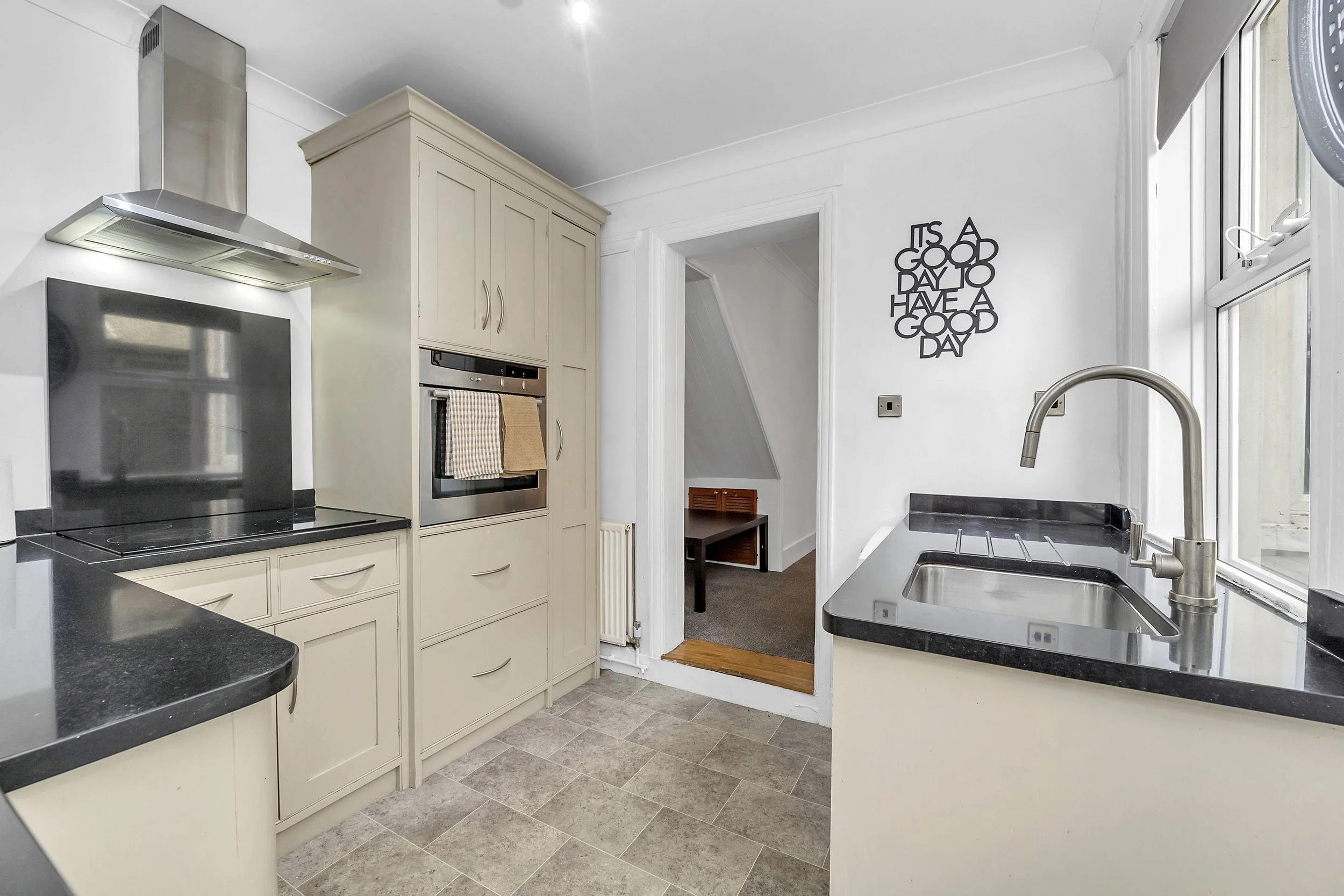 Kitchen with cream cabinets, black countertops, a stainless steel oven, and a black glass stovetop. There is a window beside a sink with a modern faucet. A wall decor reads 'It's a good day to have a good day.'