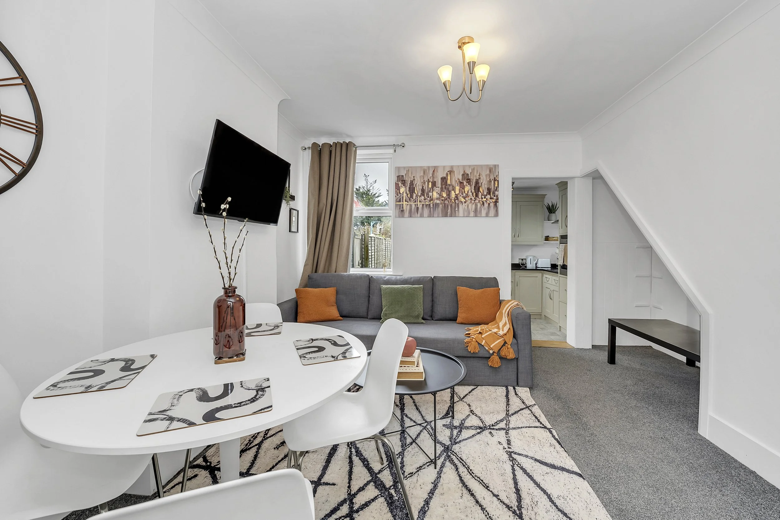 Living room with a gray sofa, orange and green pillows, a round black side table, a dining table with placemats, a wall-mounted TV, and a kitchen area in the background.