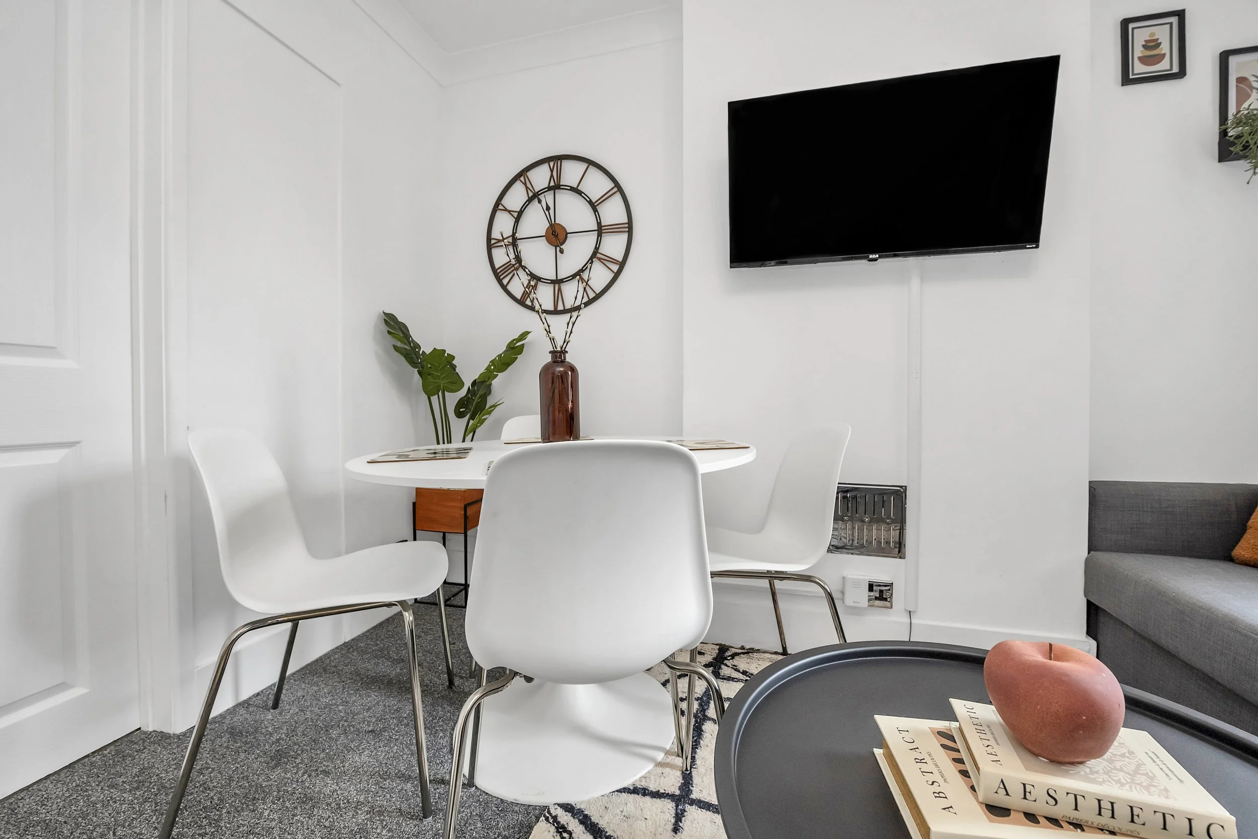 Modern living room with white walls, wall clock, wall-mounted TV, dining table with white chairs, gray sofa, and decorative items including a pink ceramic apple and books.