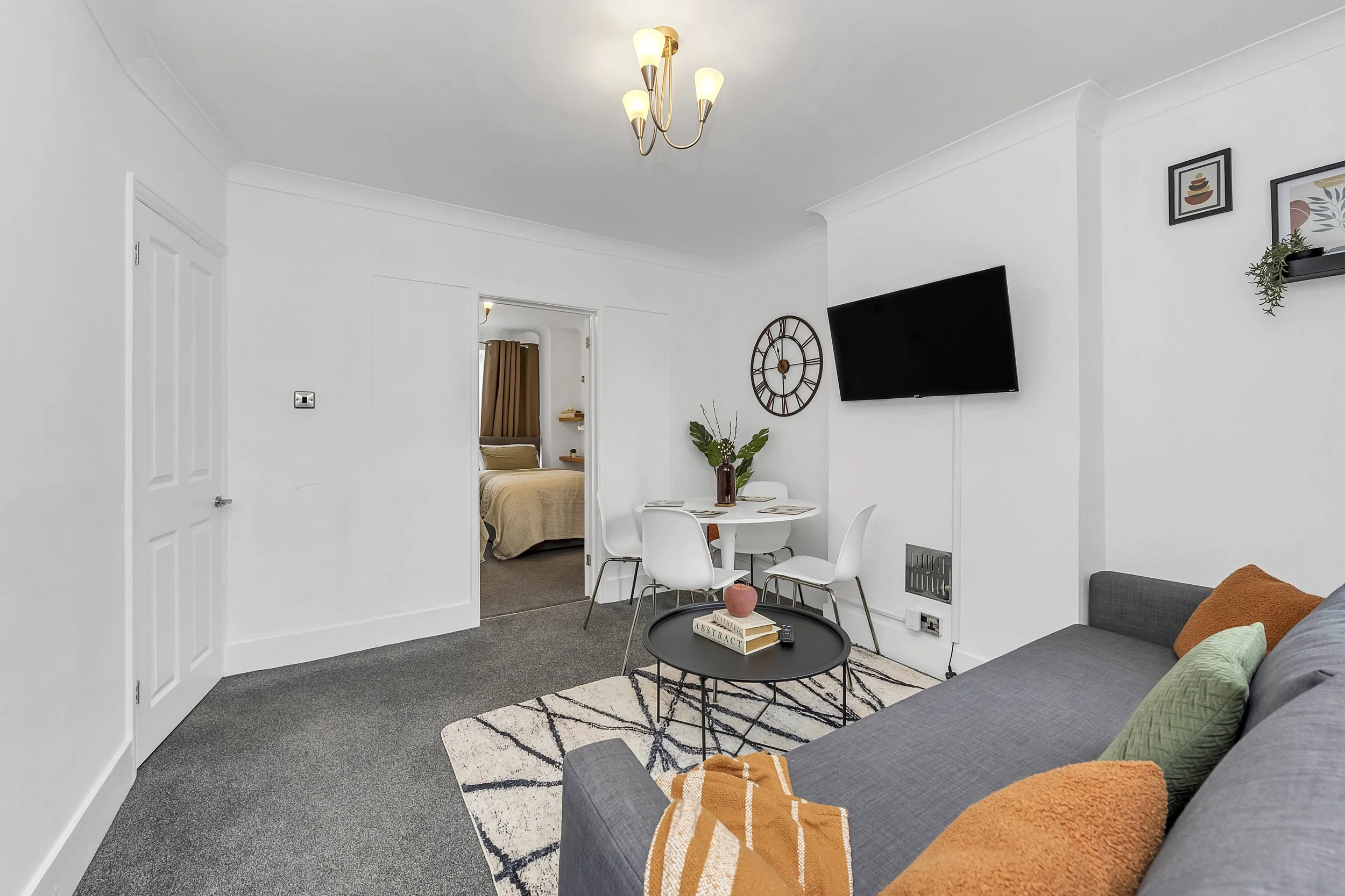 Living room with gray sofa, orange and green pillows, black round coffee table, white dining table with four chairs, wall-mounted TV, decorative wall clock, potted plant, and visible bedroom through open doorway, all in a modern decor style.