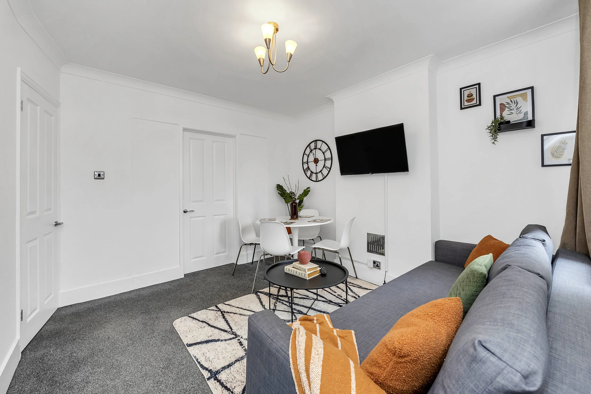 Modern living room with gray sofa, orange and green cushions, coffee table, round dining table with white chairs, wall art, wall clock, TV, textured curtain, and decorative plants.