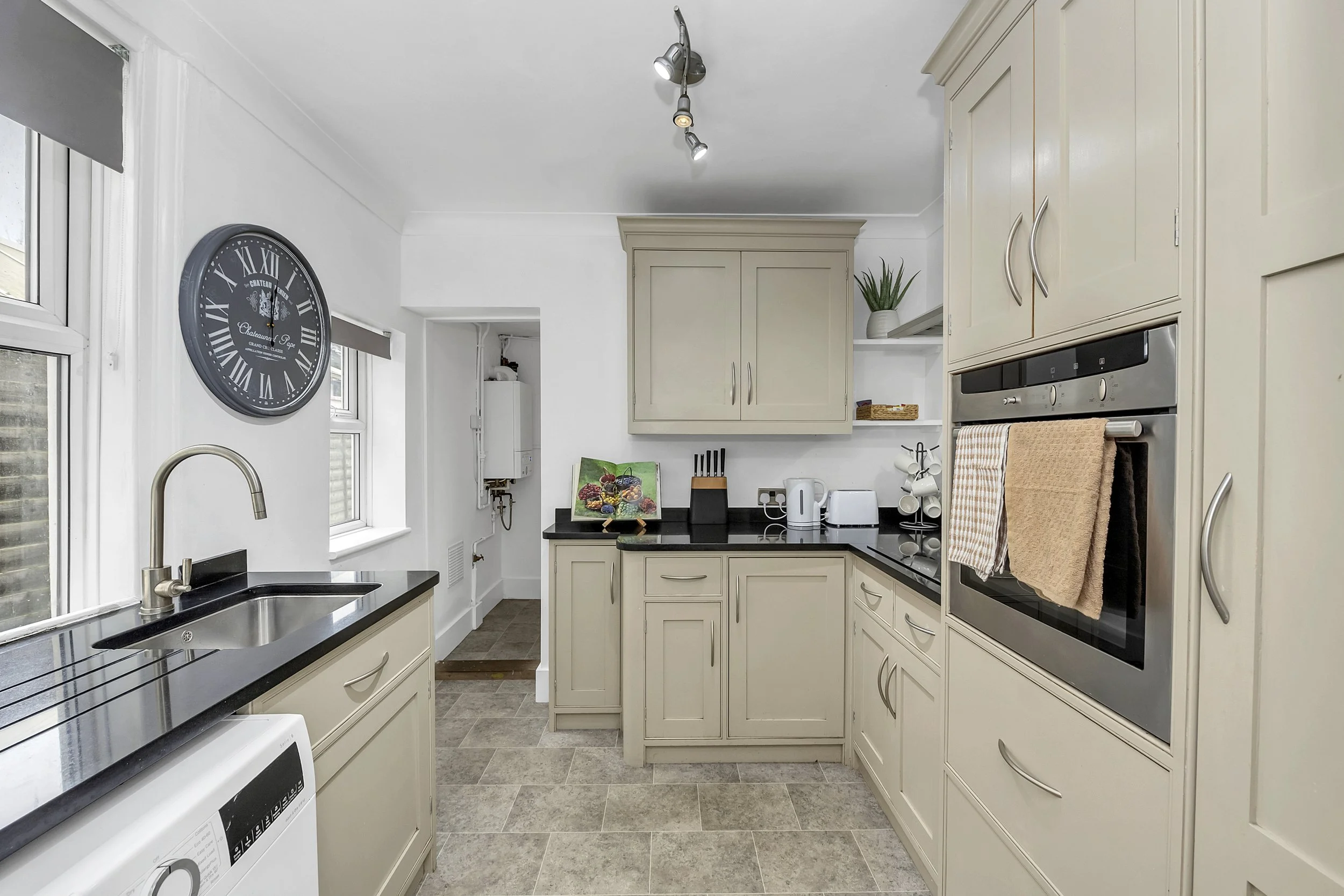 Kitchen with beige cabinets, black countertops, a window over the sink, a wall clock, and various appliances including a toaster, kettle, and oven.