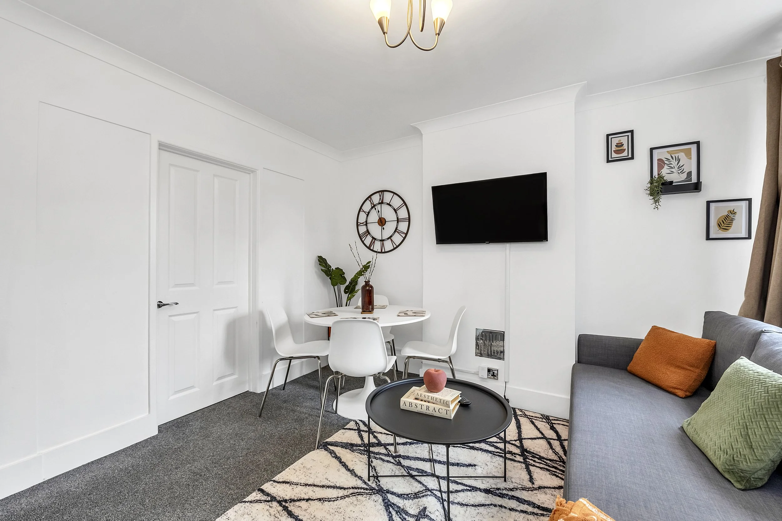 Living room with a gray sofa adorned with orange and green pillows, a black coffee table with books and a pink decorative object, a white dining table with four chairs, wall art, a large wall clock, a television mounted on the wall, and a chandelier.