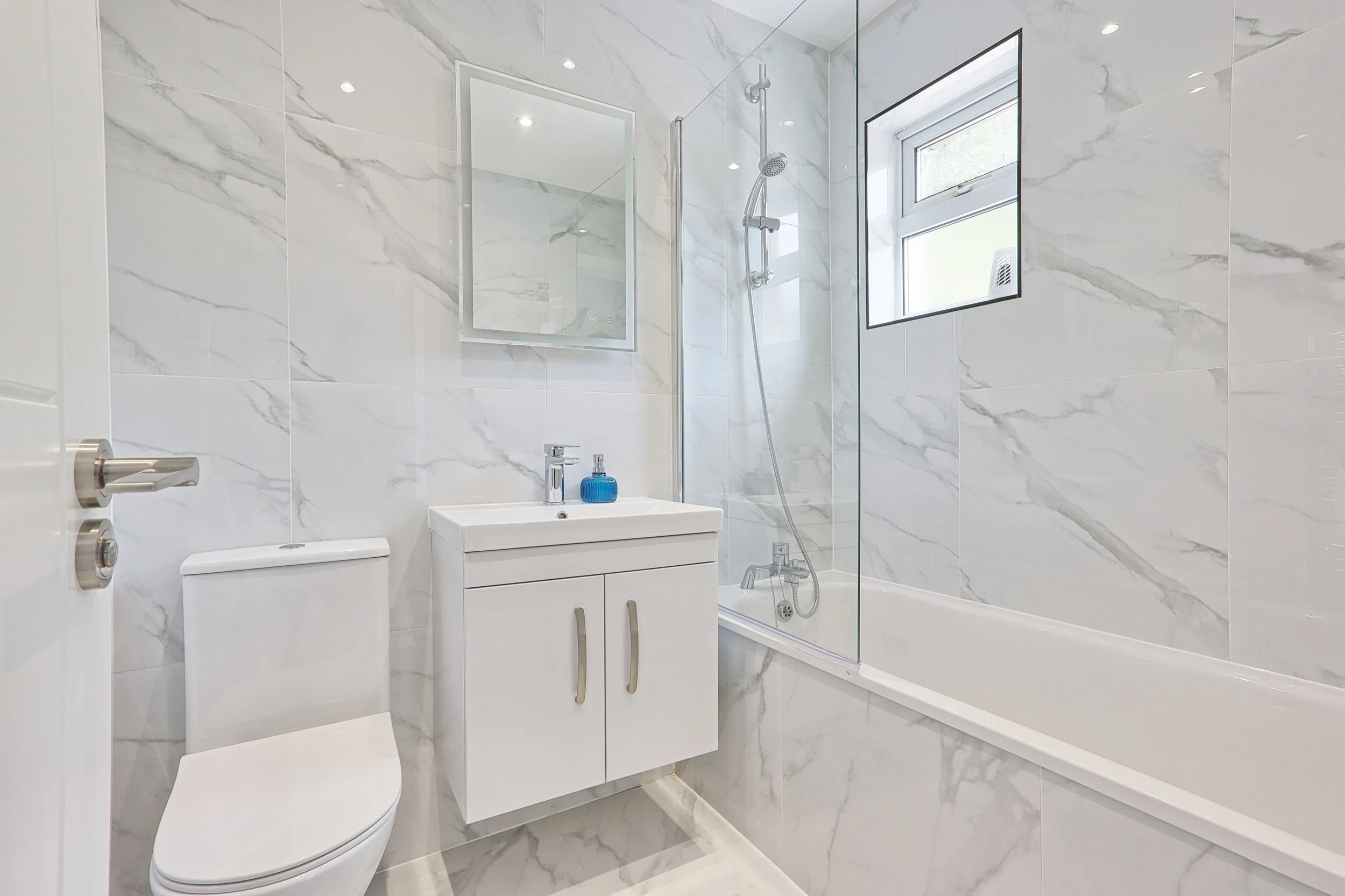 Modern bathroom with white marble walls, a toilet, a white vanity with a sink, a mirror, and a bathtub with a shower, a small window, and bright lighting.