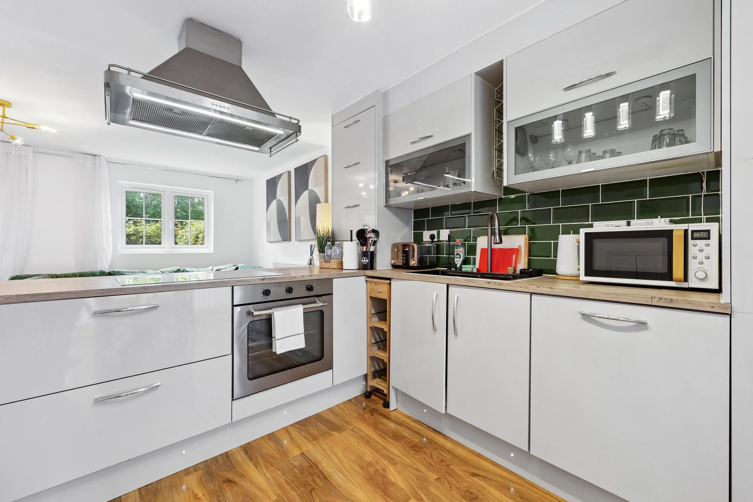 Modern kitchen with white cabinets, green tile backsplash, stainless steel oven, microwave, toaster, and kitchen utensils on a wooden countertop. There is a window with white curtains and decorative wall art in the background.