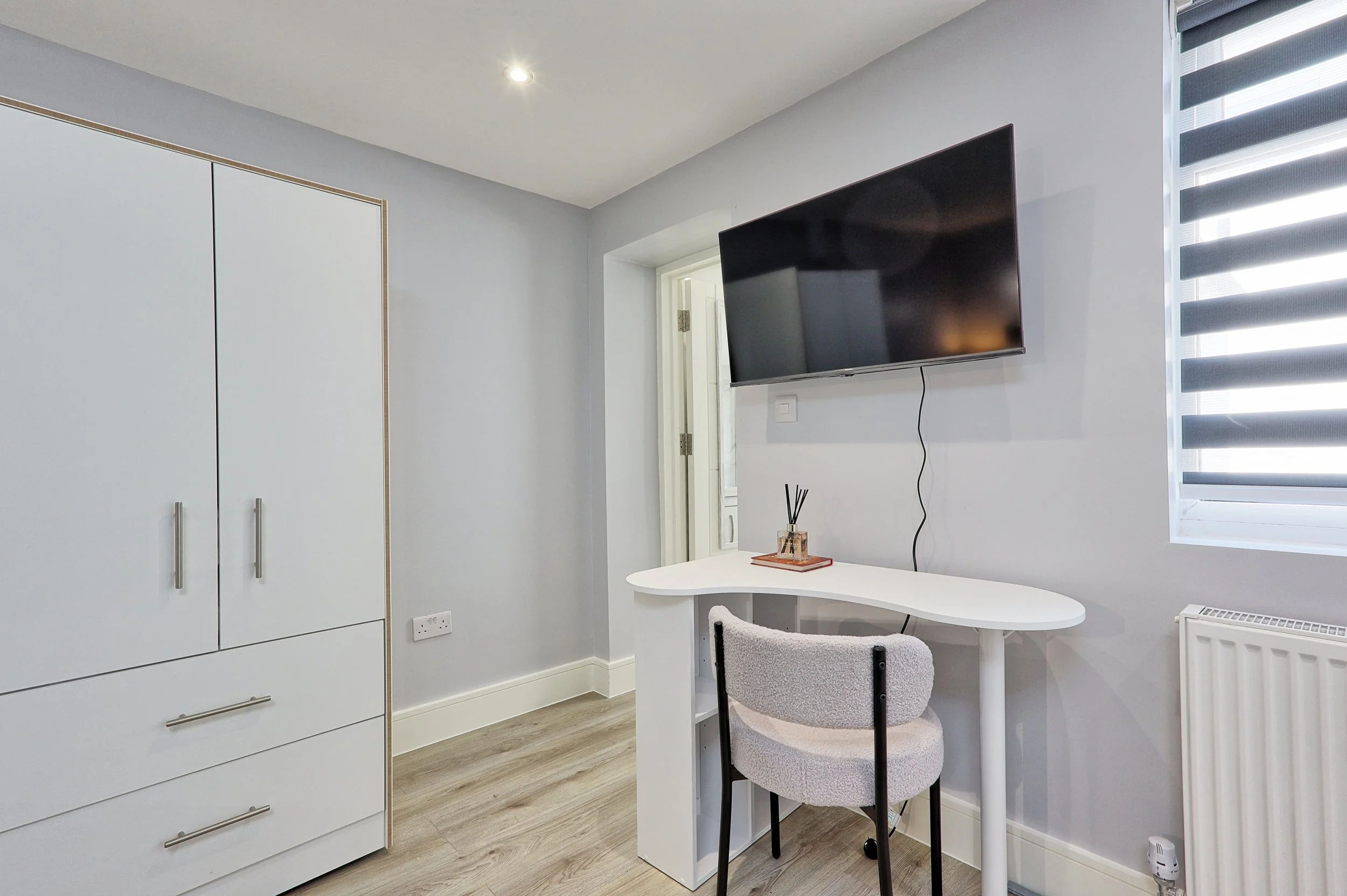 A small workspace with a white curved desk, a fluffy office chair, a wall-mounted flat-screen TV, a window with blackout blinds, a white wardrobe, and a glimpse of a door in the background.