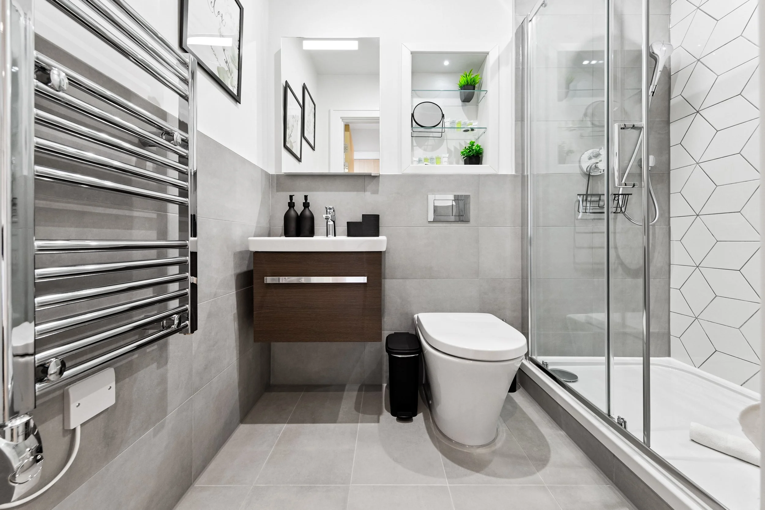 Modern bathroom with gray tiled walls and floor, white toilet, glass shower enclosure, dark wood vanity with white sink, black soap dispensers, mirror, and decorative plants on shelves.