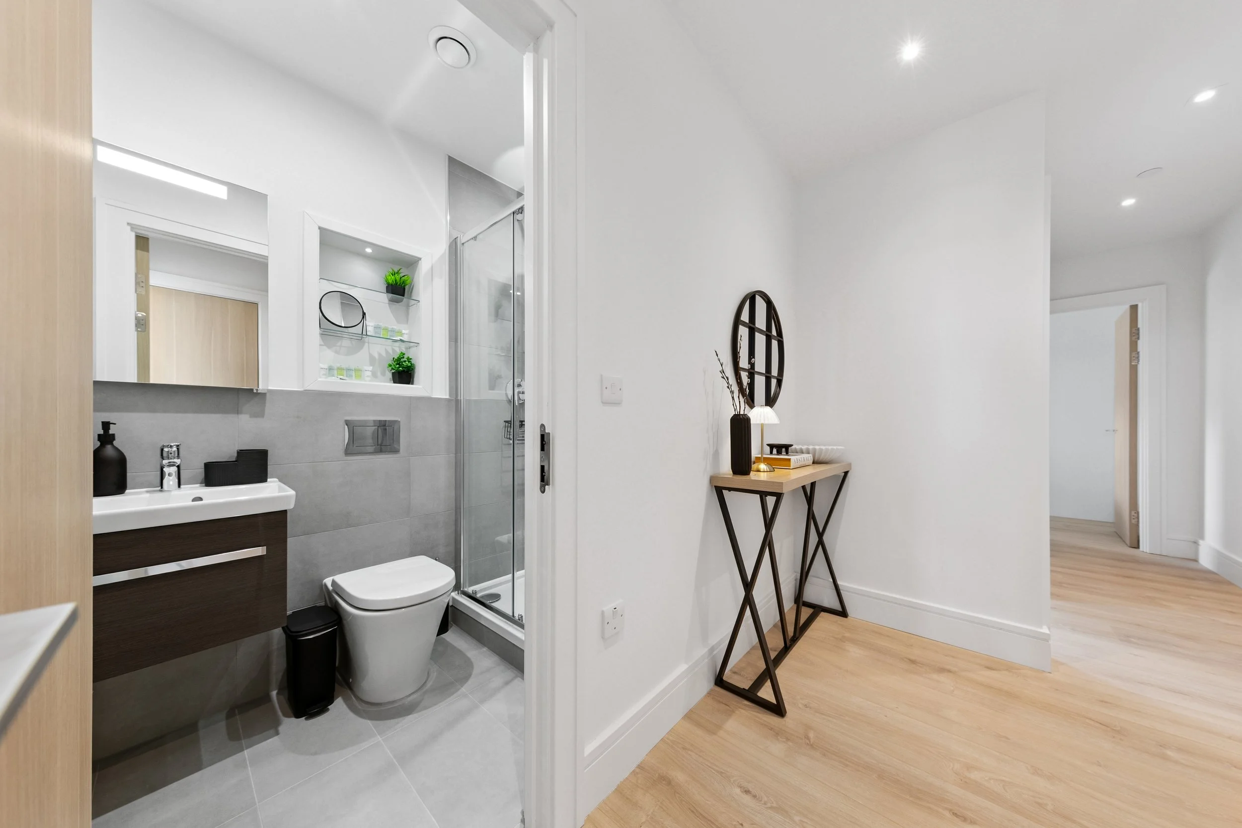 Interior view of a small bathroom with a toilet, sink, shower, and shelves with decorative items. Adjacent hallway with a console table and decor, hardwood flooring, and white walls.