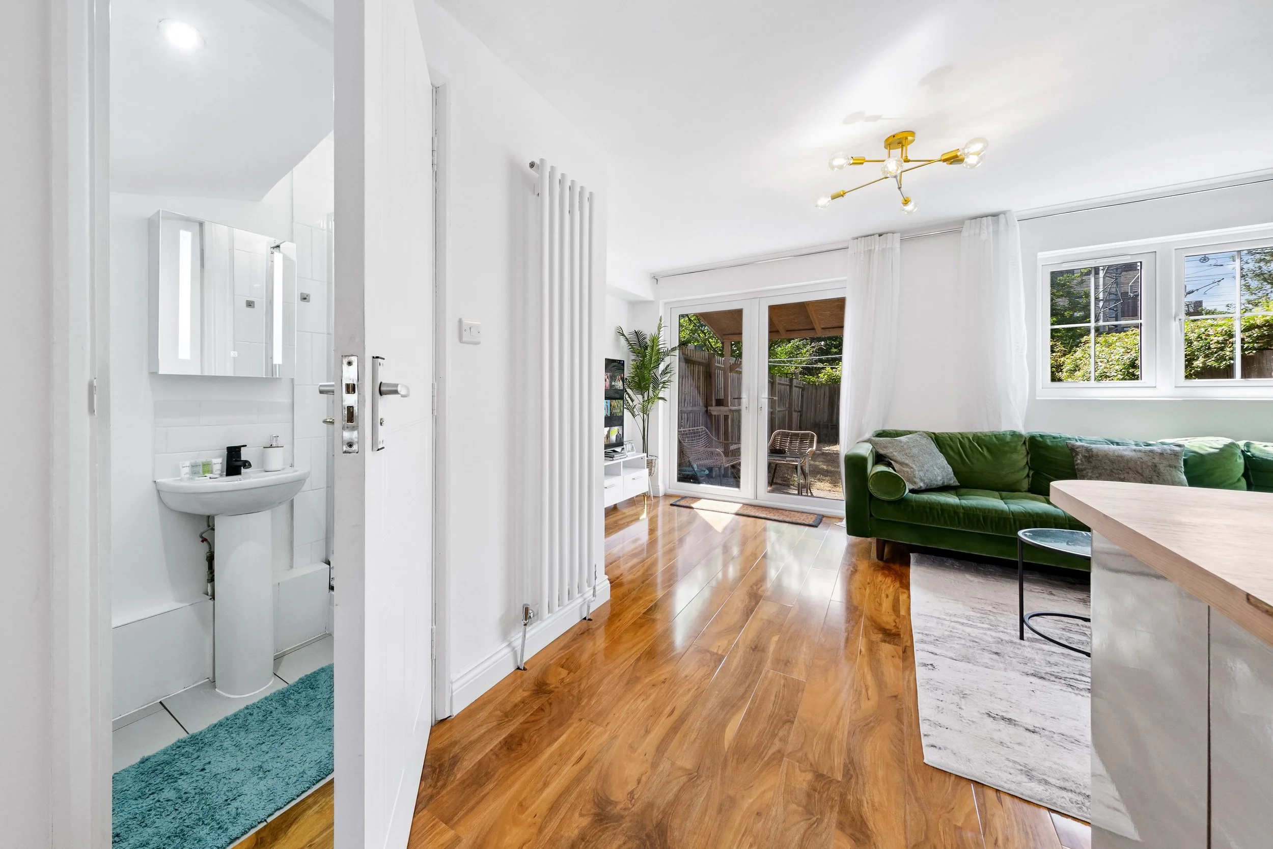 Living room with a green velvet sofa, wooden floor, sliding glass door to outdoor patio, window, modern ceiling light, and part of a kitchen with a marble countertop.