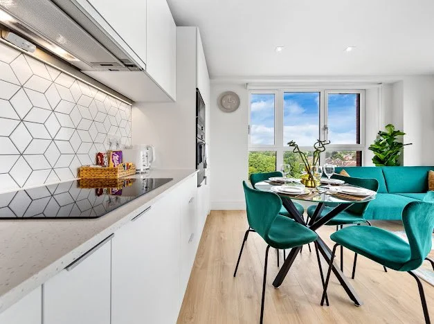 Modern kitchen and dining area with a geometric tile backsplash, white cabinets, a black induction cooktop, a round black dining table with green chairs, large window with scenery outside, and a teal sofa