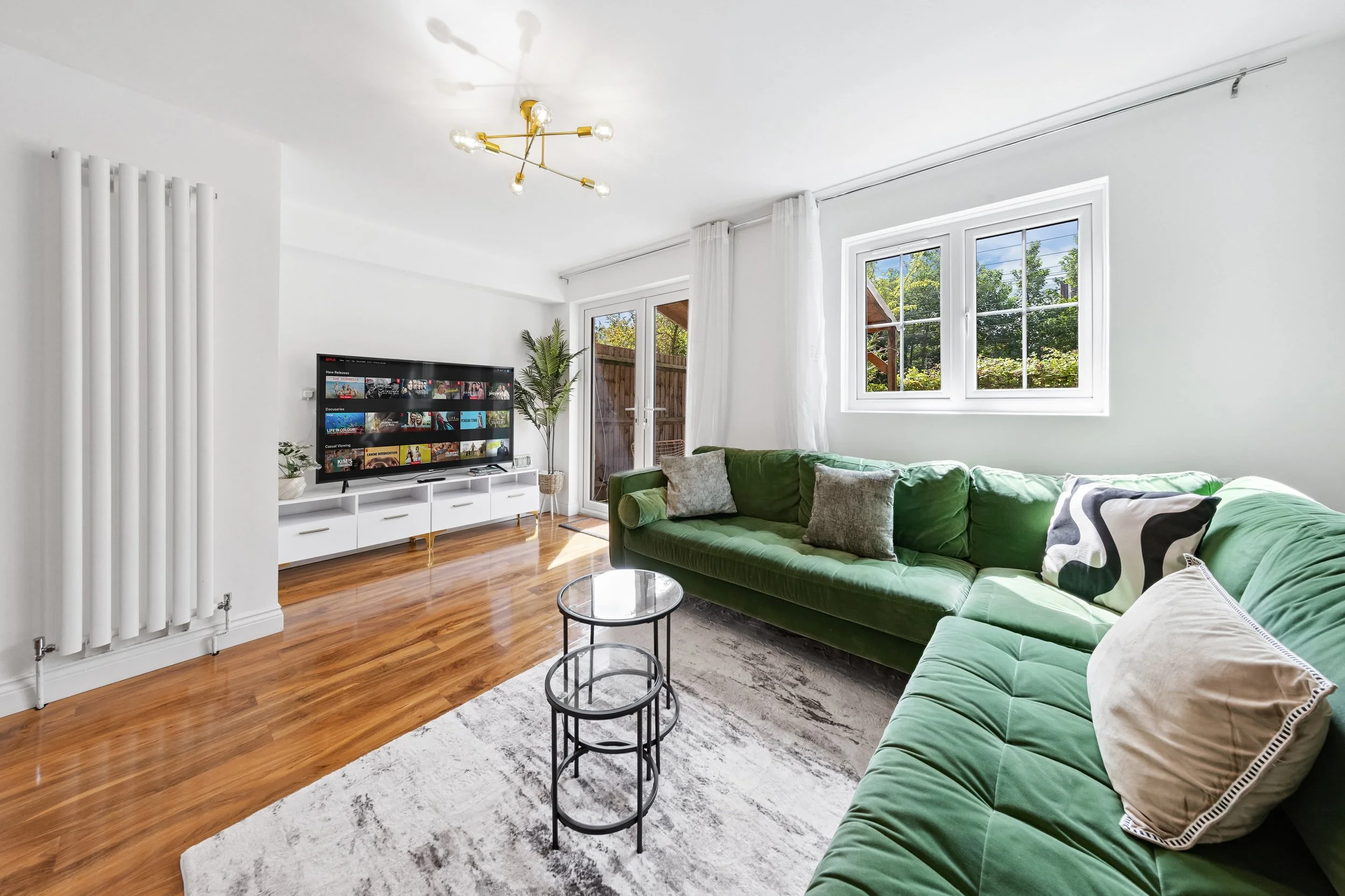 Bright living room with white walls, hardwood floors, large green sectional sofa with pillows, white tv stand with TV, potted plant, glass door, and window with white curtains.