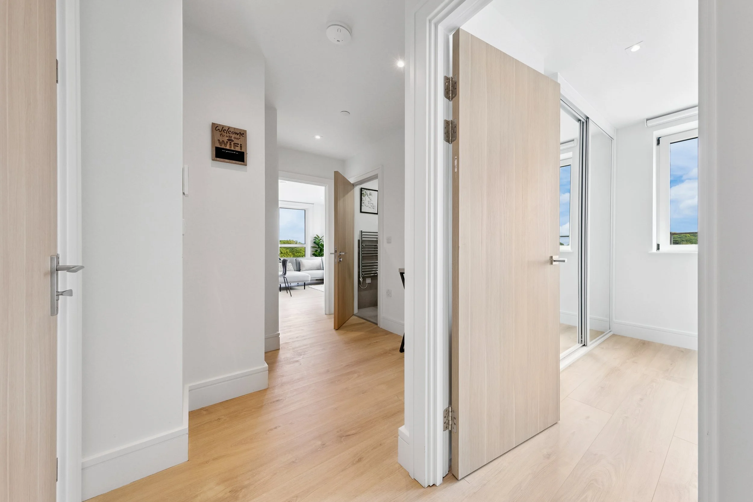 Interior view of a modern home with a hallway leading to a living room. The space features light-colored wooden floors, white walls, open doors, and windows bringing in natural light.