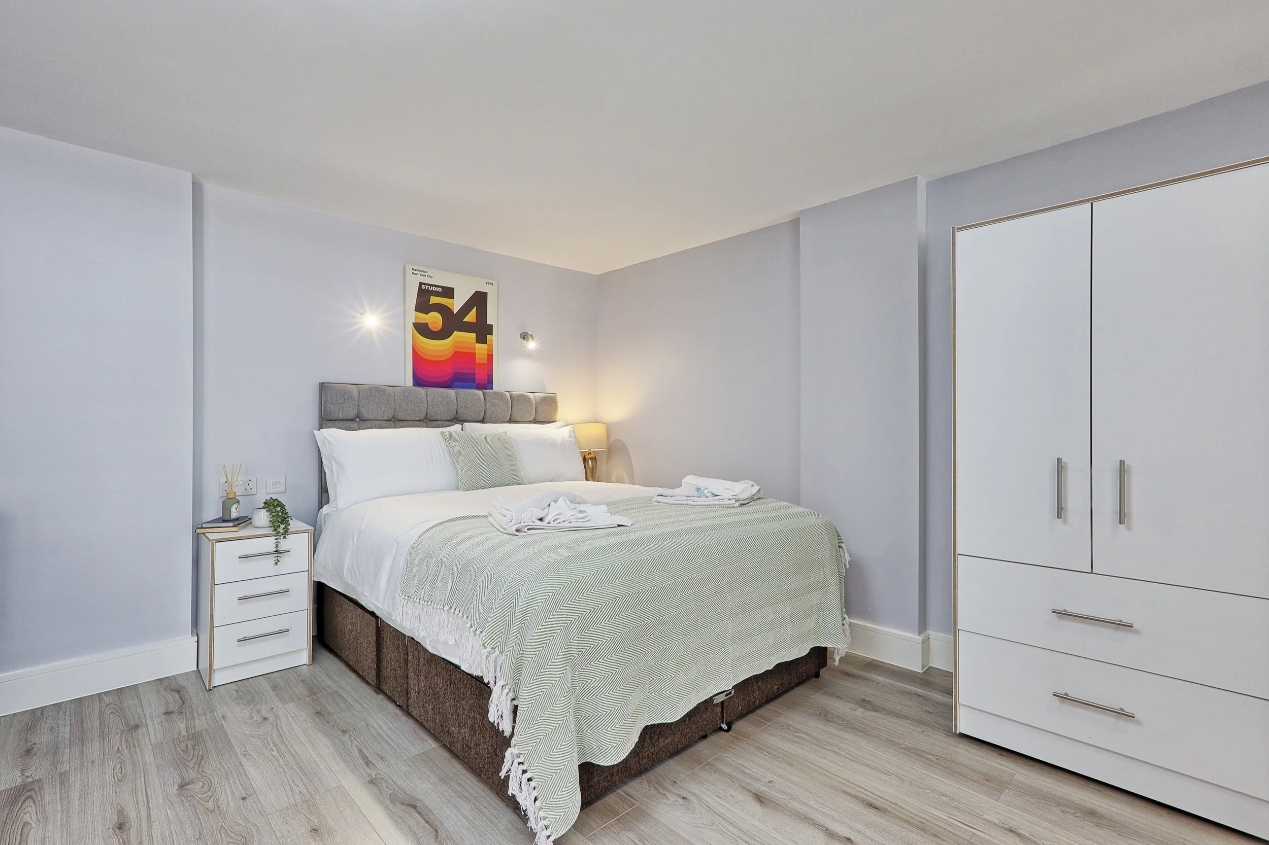 Modern bedroom with a bed, white and gray bedding, a nightstand with decor, a wardrobe, and a colorful poster above the bed.