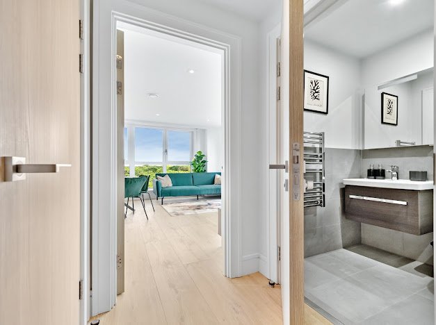 View through a doorway into a bright living room with large windows, a teal sofa, and chairs, with a small potted plant near the windows.