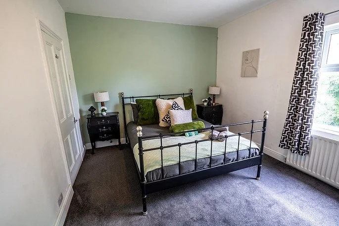 A bedroom with a black metal bed frame, green and white bedding, two black nightstands with lamps, a window with black and white patterned curtains, and a small piece of wall art.