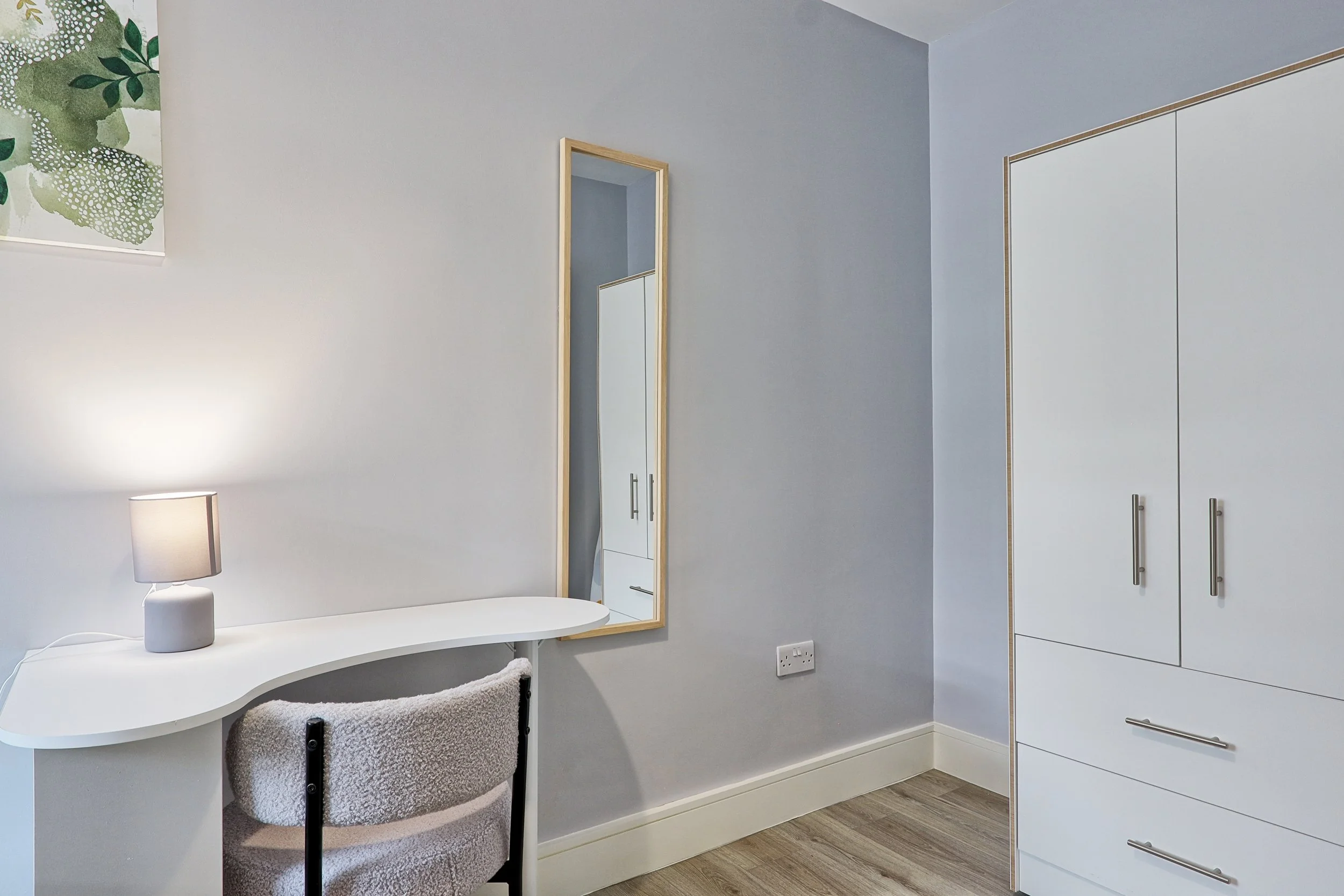 A bedroom corner with a white desk and a beige chair, a small table lamp, a full-length mirror, a white wardrobe, and a floral wall art