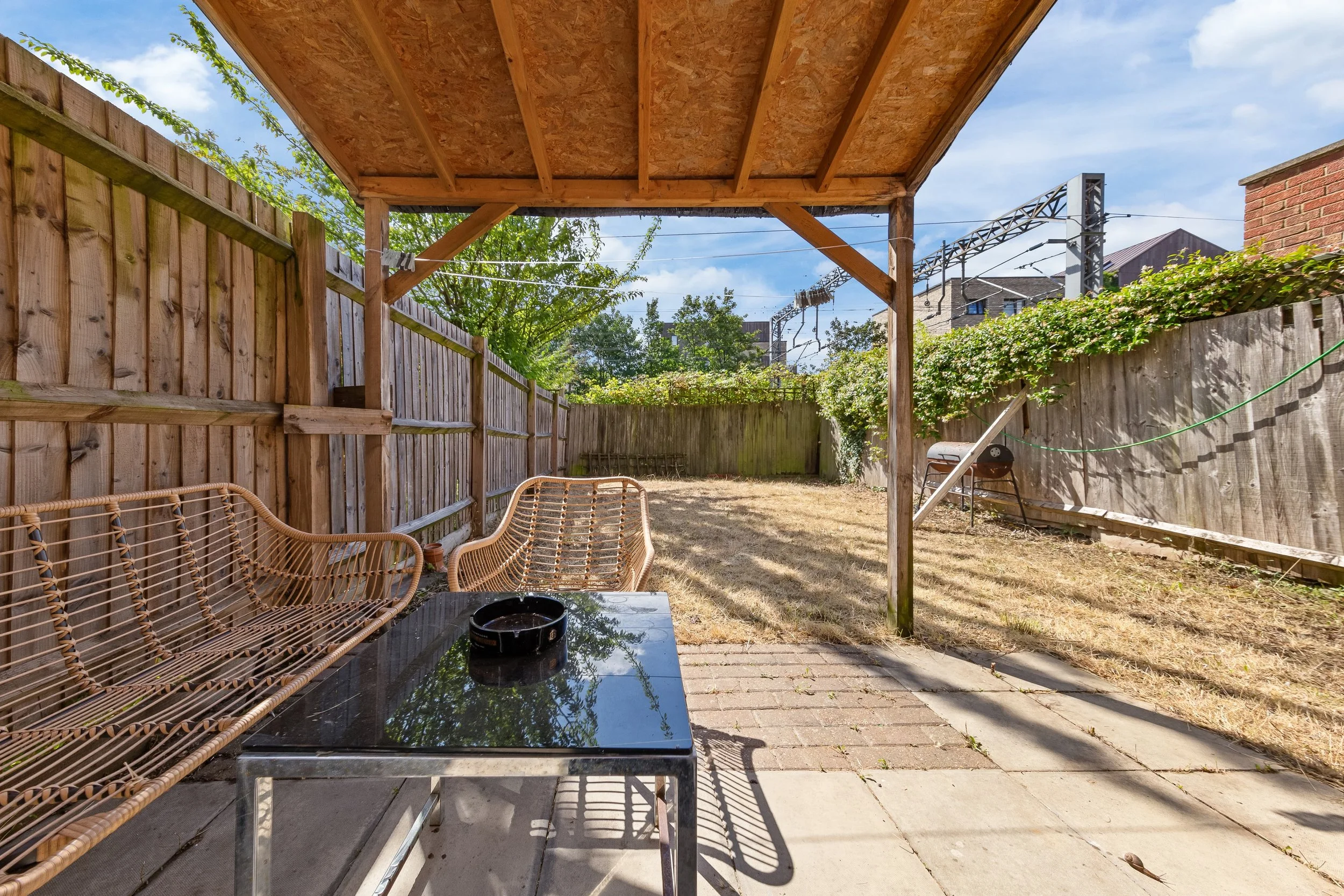 Backyard patio area with a shaded wooden pergola, wicker seating, a glass table with a black ashtray, and a grassy yard enclosed by wooden fencing and a chain-link fence. There are electric lines and a building across the sky.