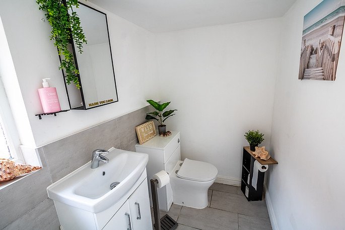 Small bathroom with white walls, a white sink with a cabinet, a white toilet, and decorative elements including plants, a framed sign, and seaside-themed artwork.