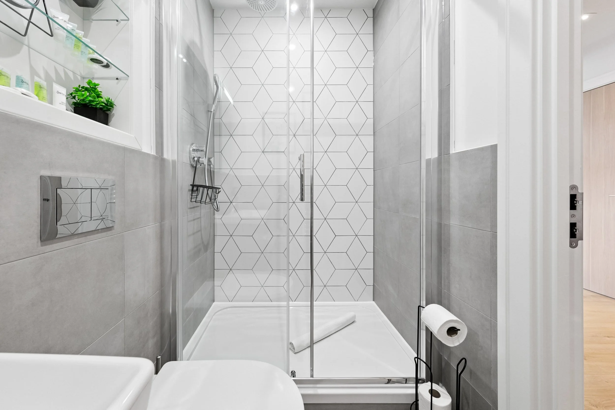 Modern bathroom with gray tiled walls, a glass shower enclosure, and a white toilet with a black toilet paper stand.