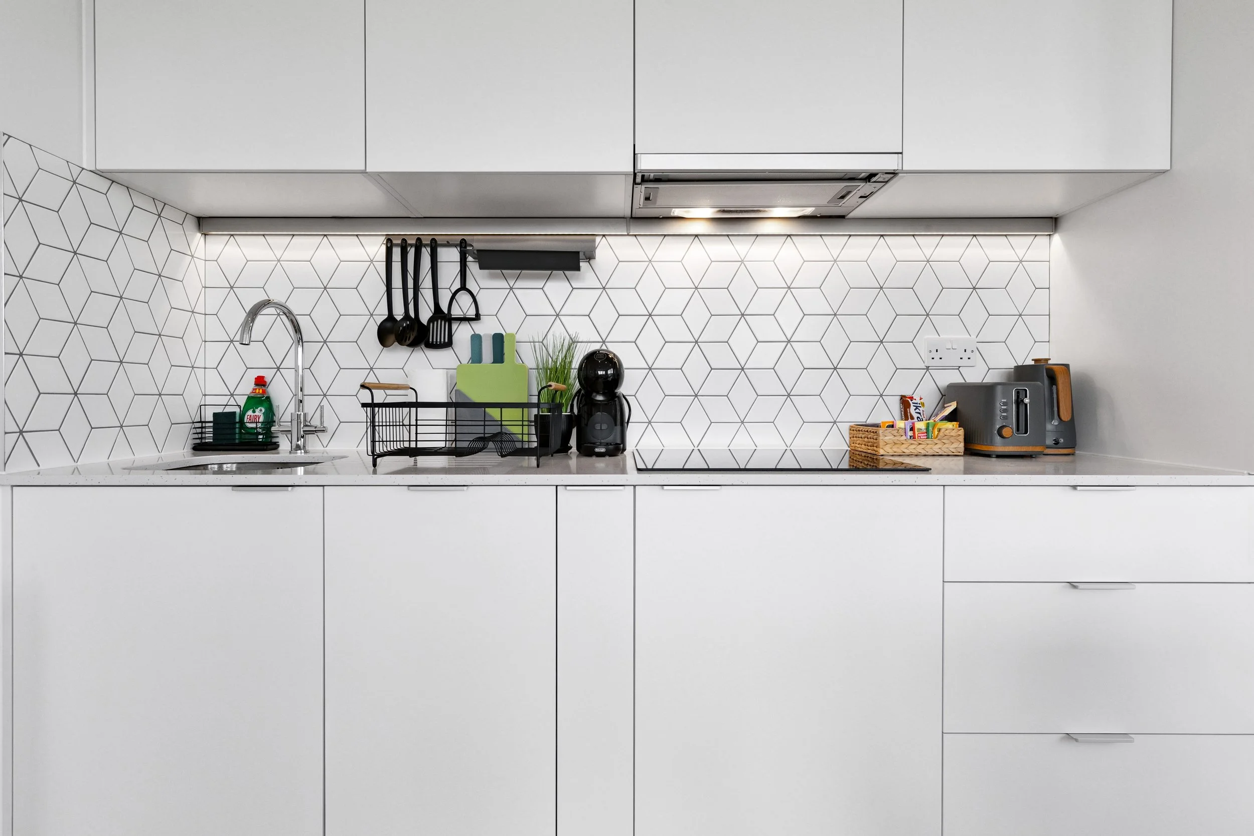 Modern minimalist kitchen with white cabinets, geometric patterned white backsplash, black hanging utensils, green cutting board, black coffee machine, small wicker basket of snacks, and gray toaster on countertop.