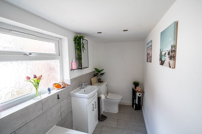 A bathroom with a large window, a white sink, a toilet, and decorative plants and artwork on the walls.