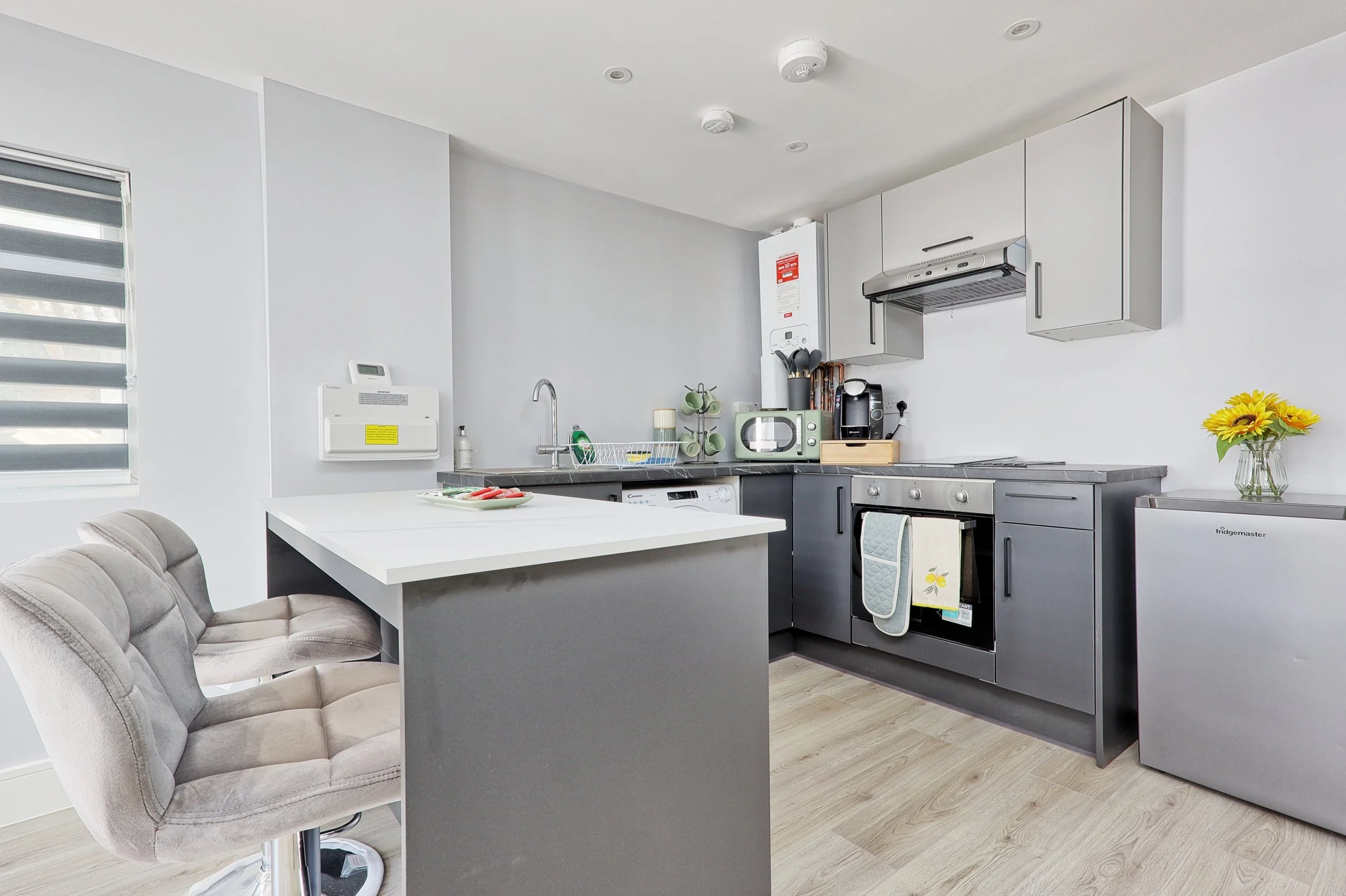 Modern kitchen with gray and white cabinets, stainless steel appliances, a small white breakfast bar, two upholstered chairs, and a vase of yellow sunflowers.