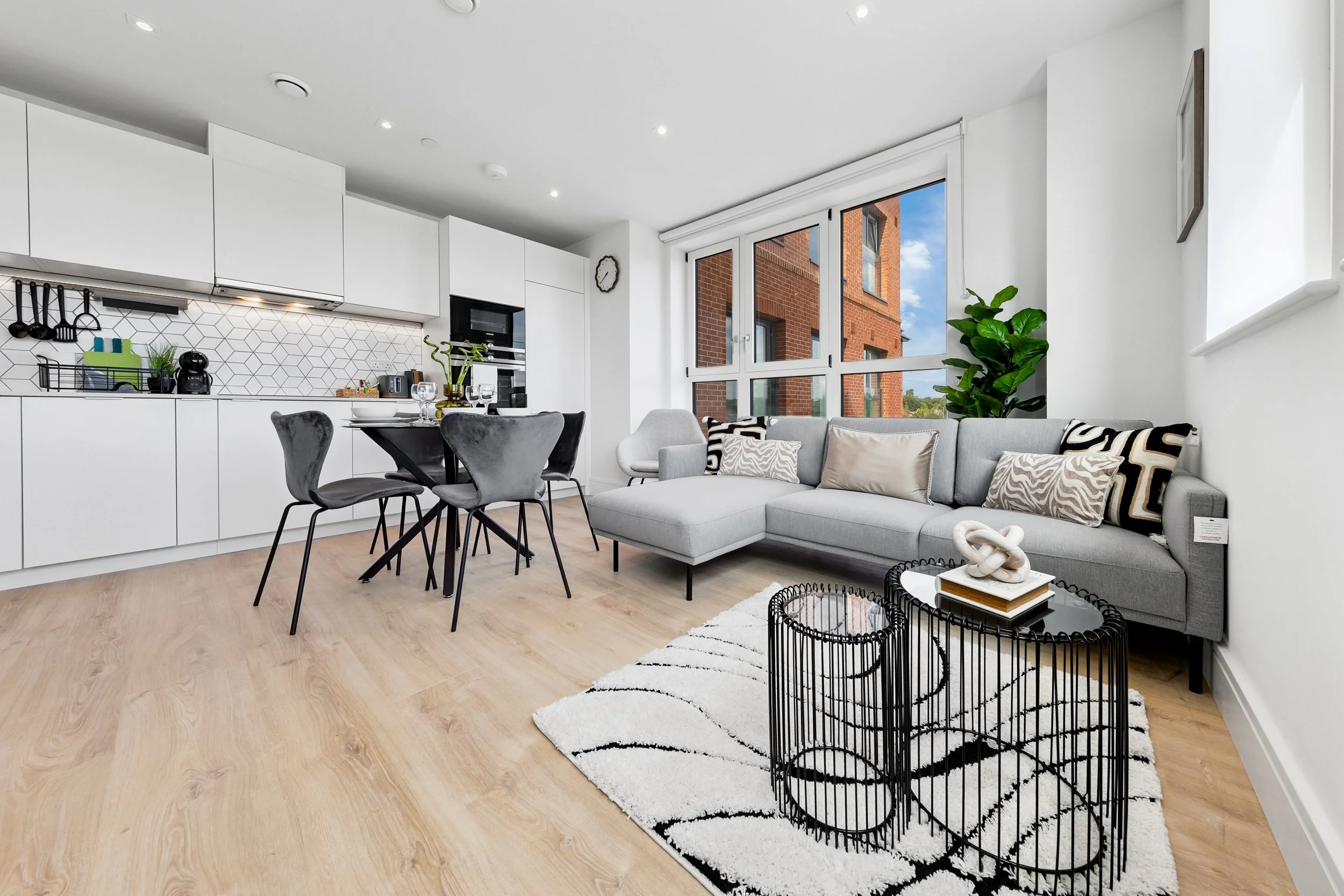 Open-concept living room and kitchen with white cabinets, gray sofa, black round tables, and large windows showing a brick building outside.