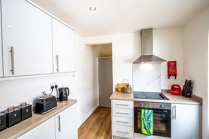 Kitchen with white cabinets, black toaster, electric kettle, countertop, oven, stovetop, range hood, red fire safety equipment, and a colorful dish towel.