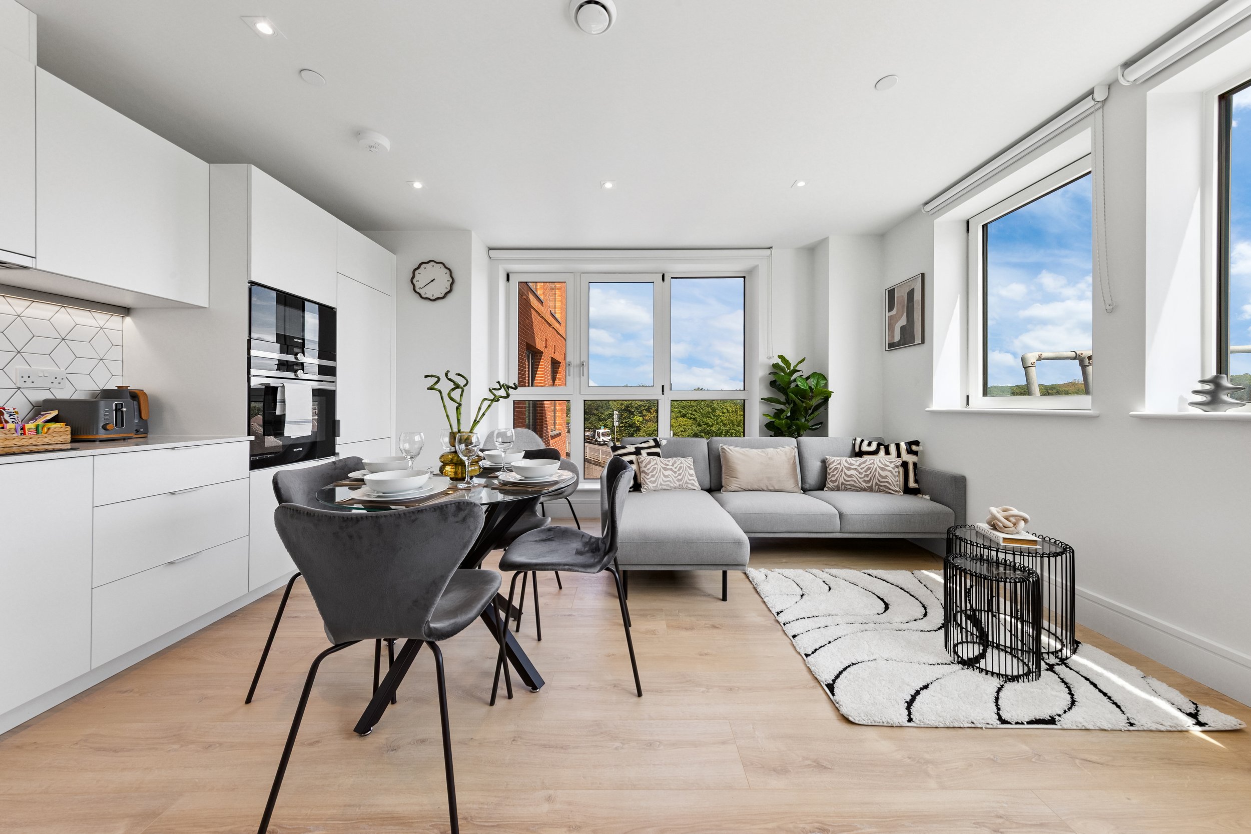 Modern open-concept living room and kitchen with white cabinetry, black dining table with chairs, gray sectional sofa with throw pillows, large windows showing green trees and blue sky, hardwood floors, and contemporary decor.