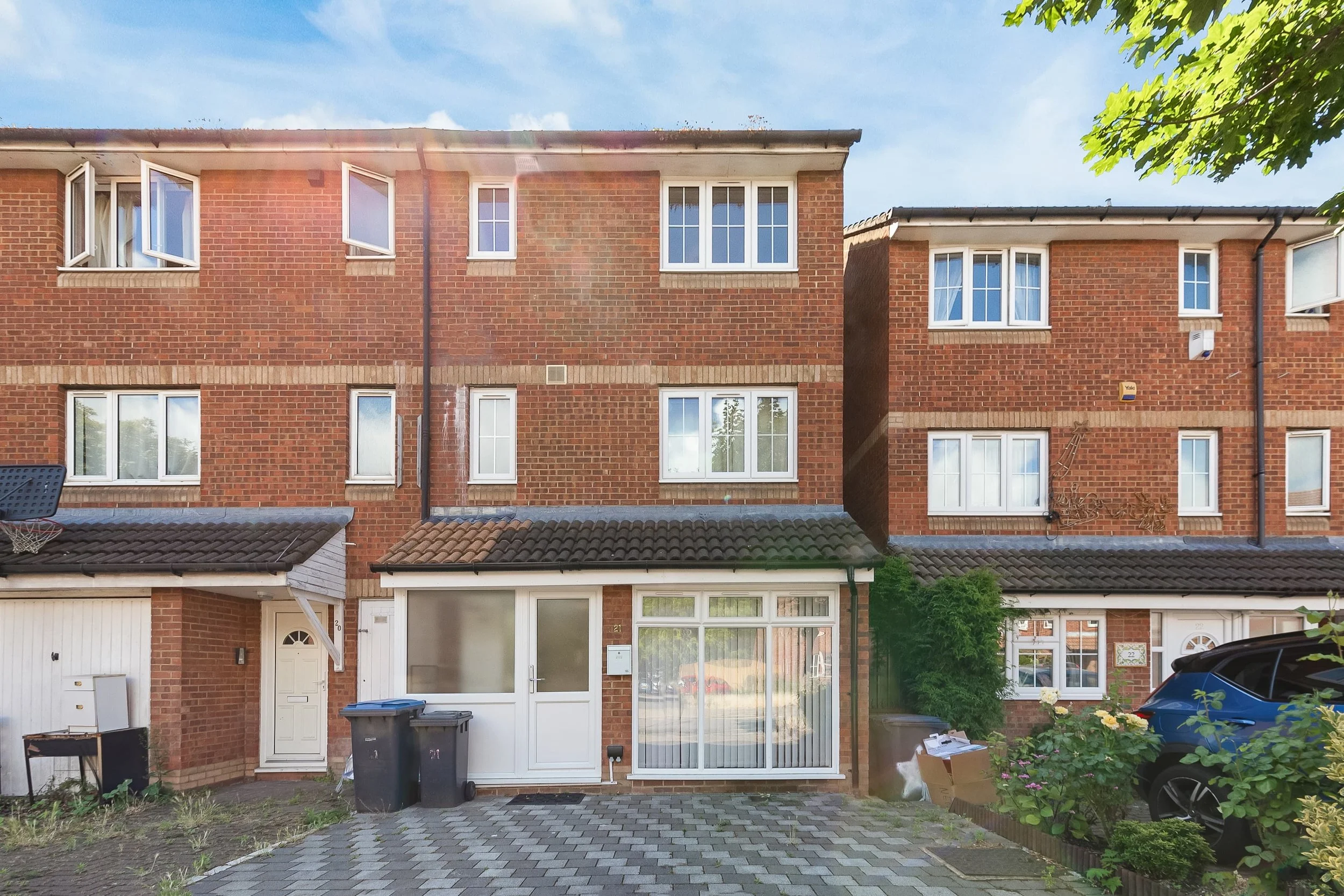 Front view of three-story brick townhouse with bay window, front door, and driveway, with greenery and parked car.