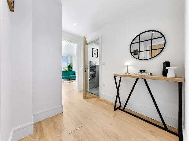 Entryway with a black round wall mirror, wooden console table with decorative items, and a partially open door leading to a bathroom. Visible part of a living room with a teal sofa.