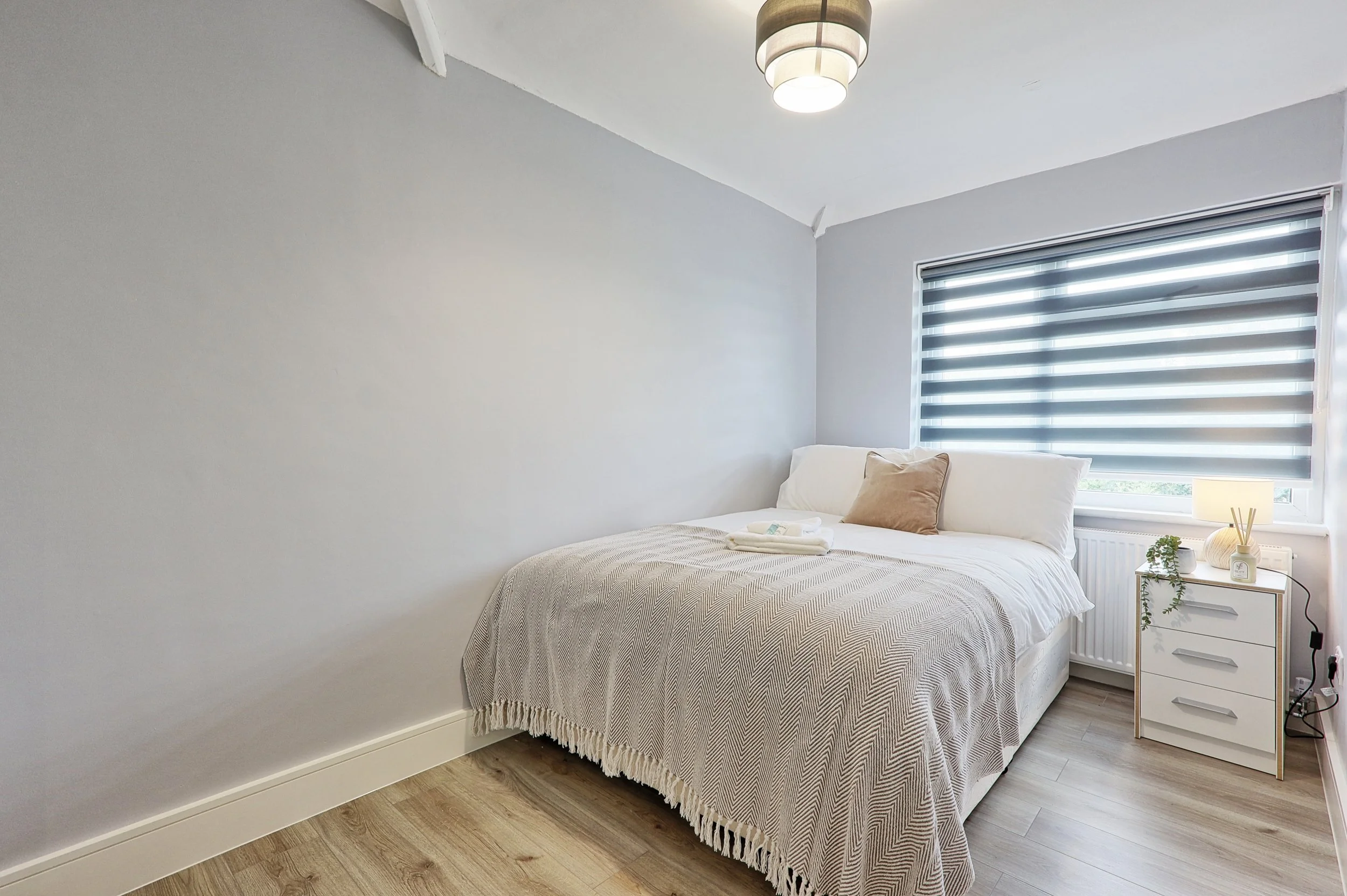 Photo of a cozy bedroom with a bed, white bedding, pillows, a small bedside table with decor, a window with striped blackout blinds, and wooden flooring.
