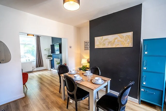 Dining area with a wooden table set for four, dark chairs, a dark accent wall with a light-colored painting, blue lockers, and a living room with a window and a television in the background.