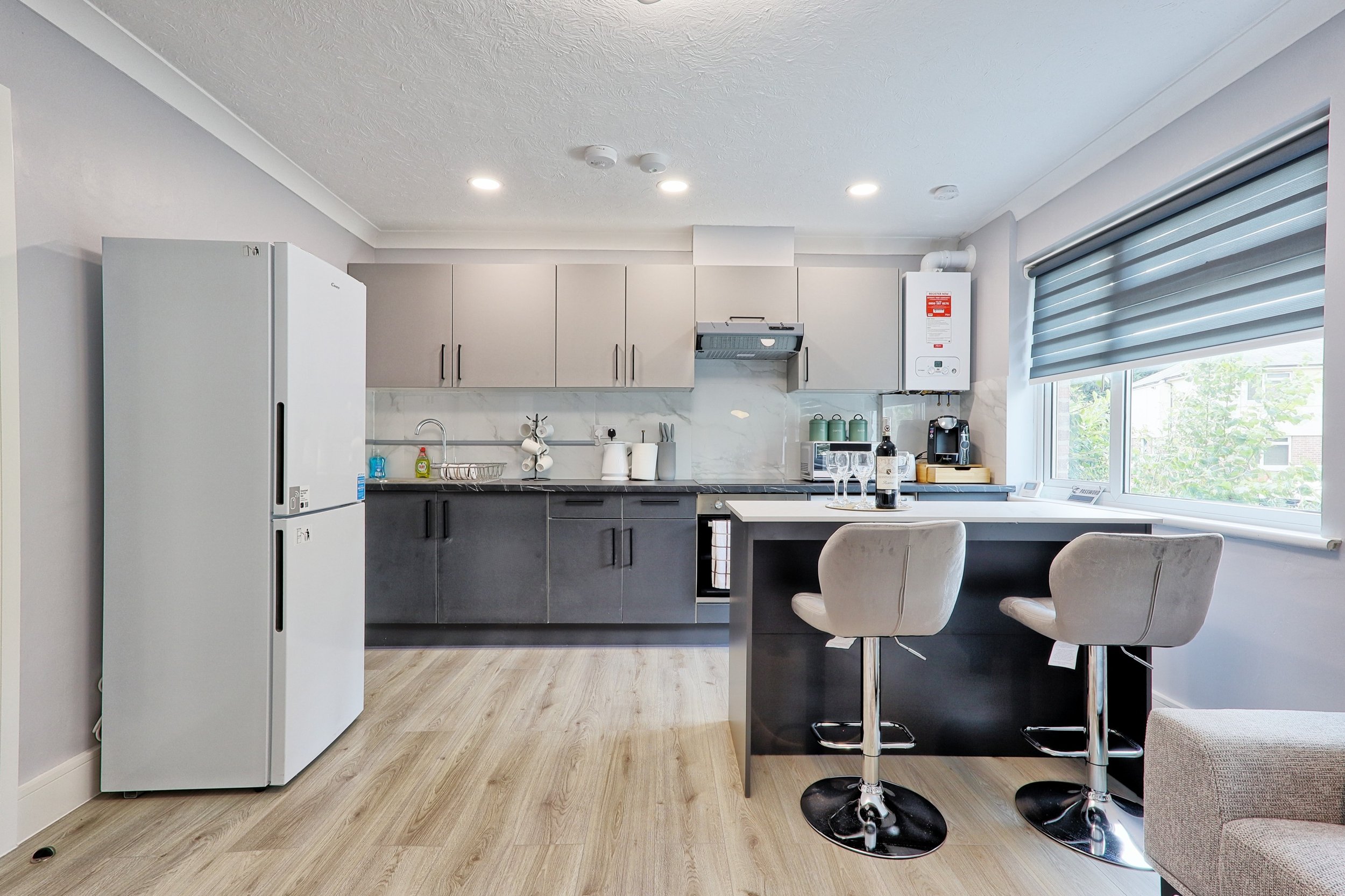 Modern kitchen with gray cabinets, a white refrigerator, a small window with a roller blind, and a breakfast bar with two stools.