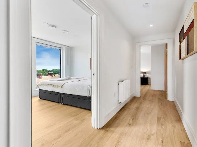 Empty hallway with light wood flooring, white walls, and a doorway leading to a bedroom with a large window, bed, and outdoor view