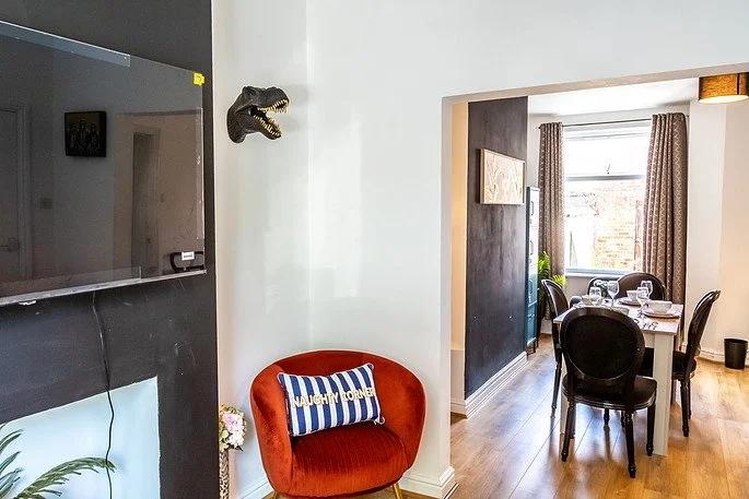 Living room with a wall-mounted television, a red velvet chair with a striped cushion, a dinosaur head wall decoration, and a view into the dining area with a table set for four and curtains in the background.