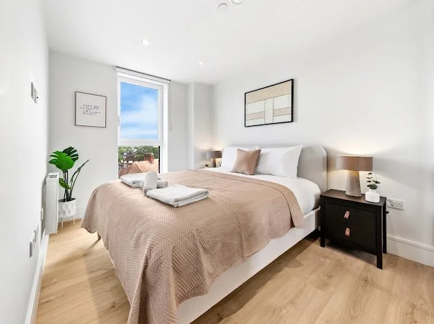 A bright bedroom with a large window showing a view of rooftops and a blue sky, a white bed with beige and pink pillows, a beige quilt, black nightstands with lamps, a small potted plant, and framed wall art.