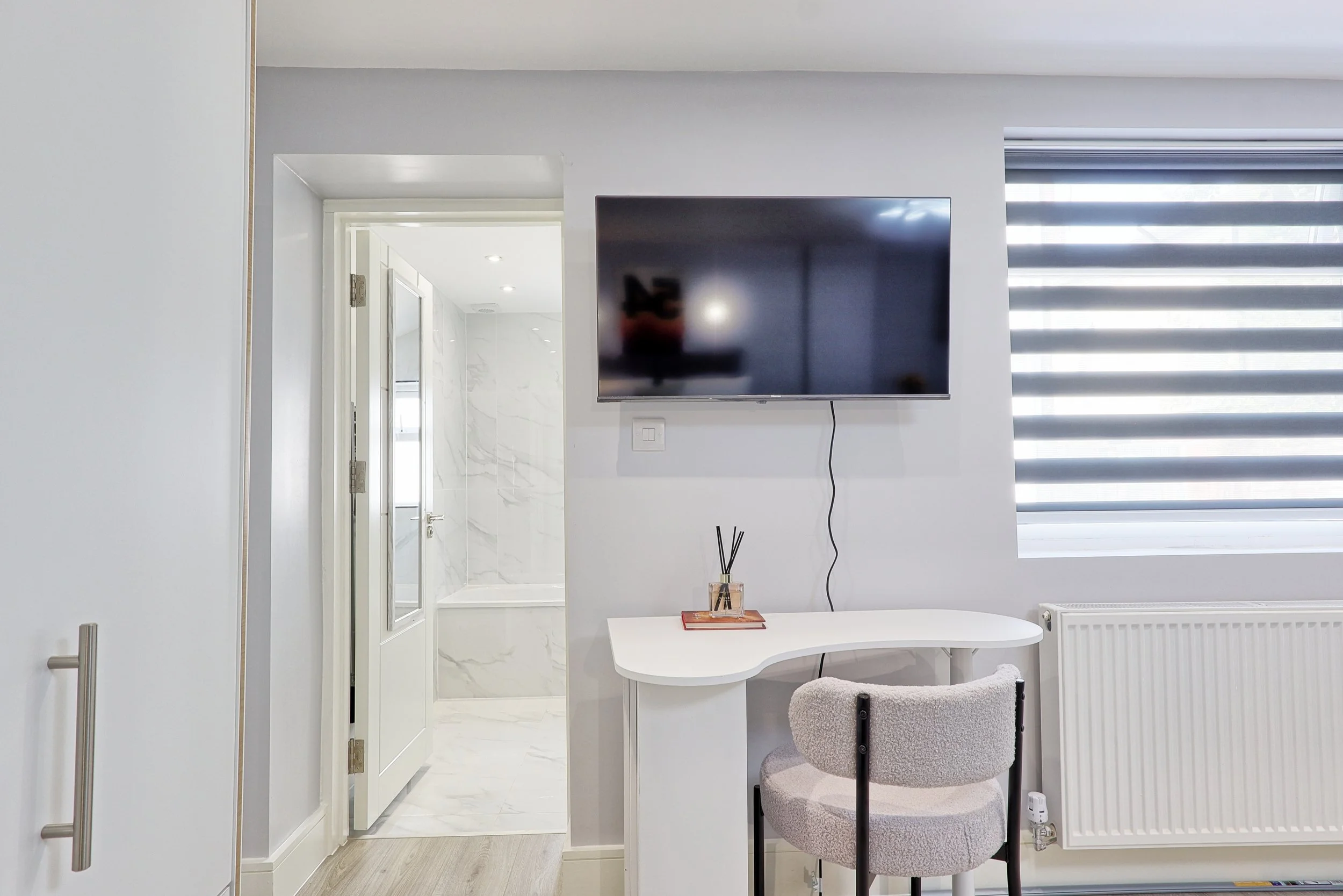 A modern interior room with white walls, a wall-mounted flat-screen TV, a small white desk with a beige cushioned chair, and a window with horizontal black and white blinds. There's a bathroom visible through a doorway, featuring white marble walls and a glass shower enclosure.