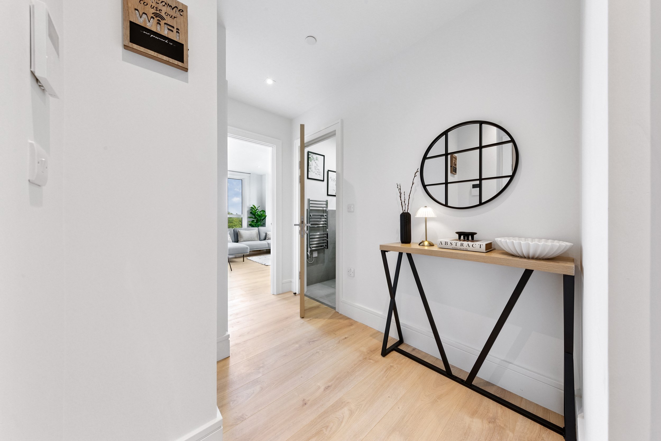 Modern hallway with a console table, decorative mirror, lamp, and vase, leading to a bright living room with large windows and gray sofa.