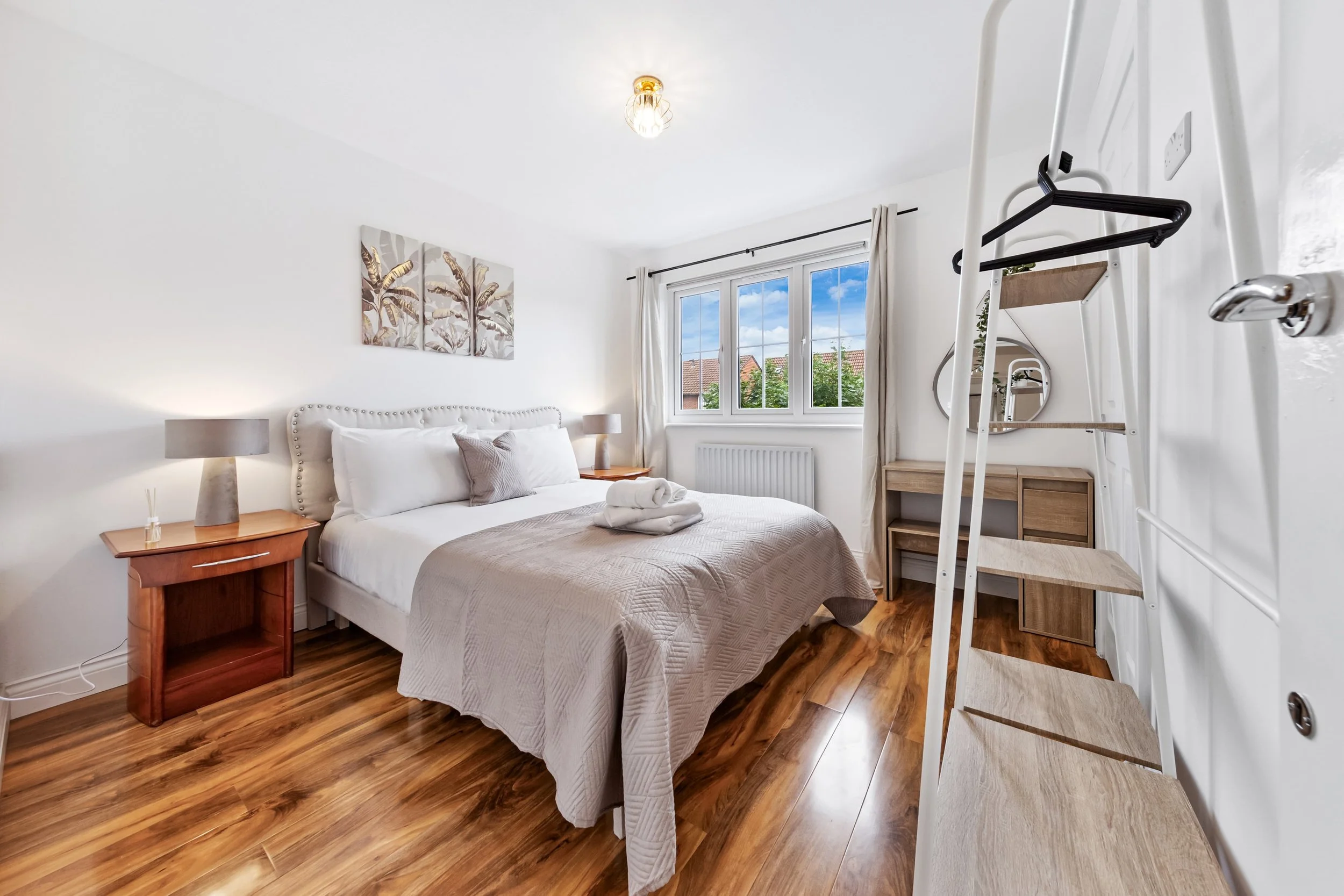 A clean and cozy bedroom with a large bed, wooden nightstands with lamps, framed wall art, a window with view of a suburban neighborhood, and a ladder-style shelf with a mirror and small items.