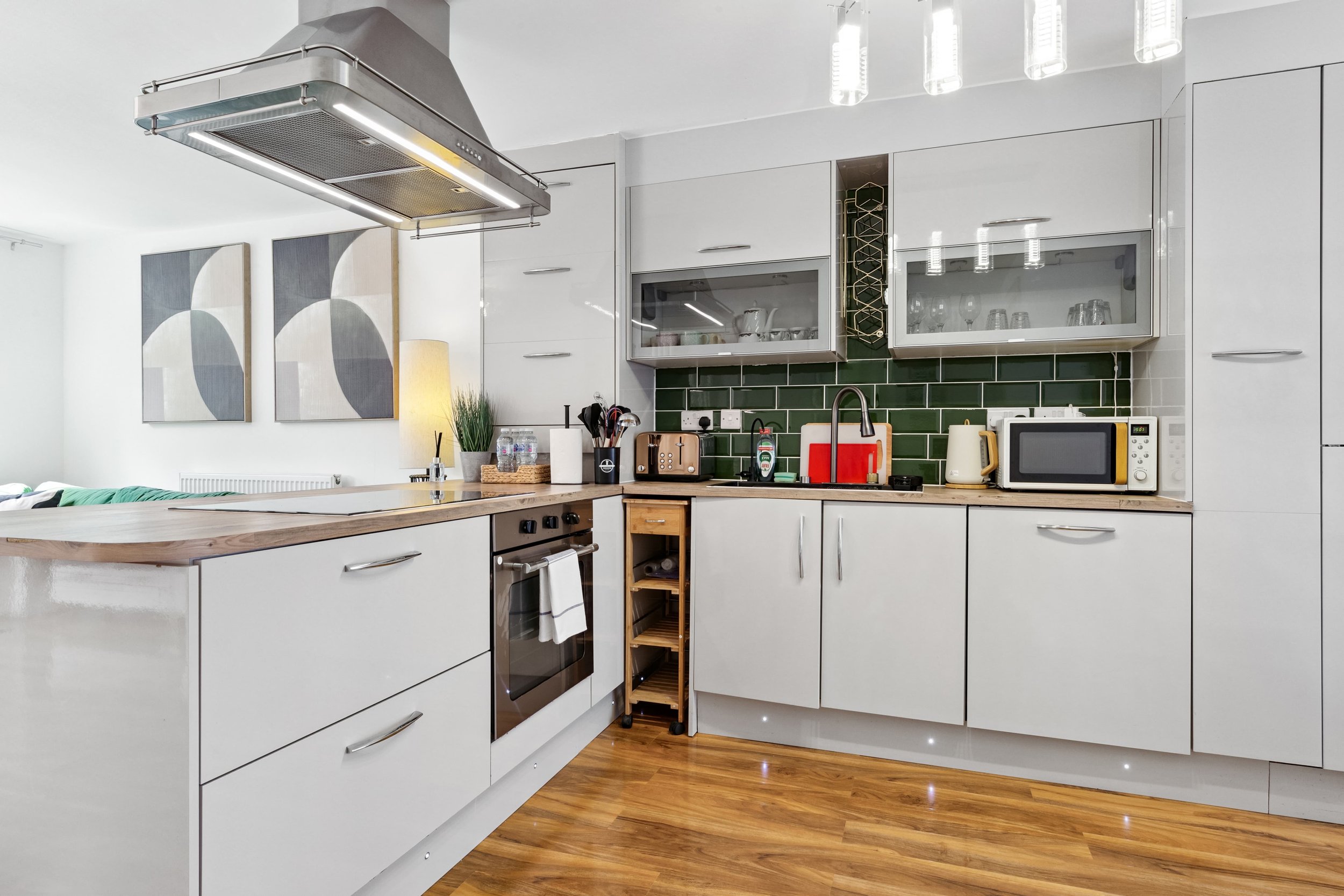 Modern kitchen with white cabinets, wooden countertop, green subway tile backsplash, microwave, toaster, kettle, and various kitchen utensils. Wooden floor and abstract wall art in the background.