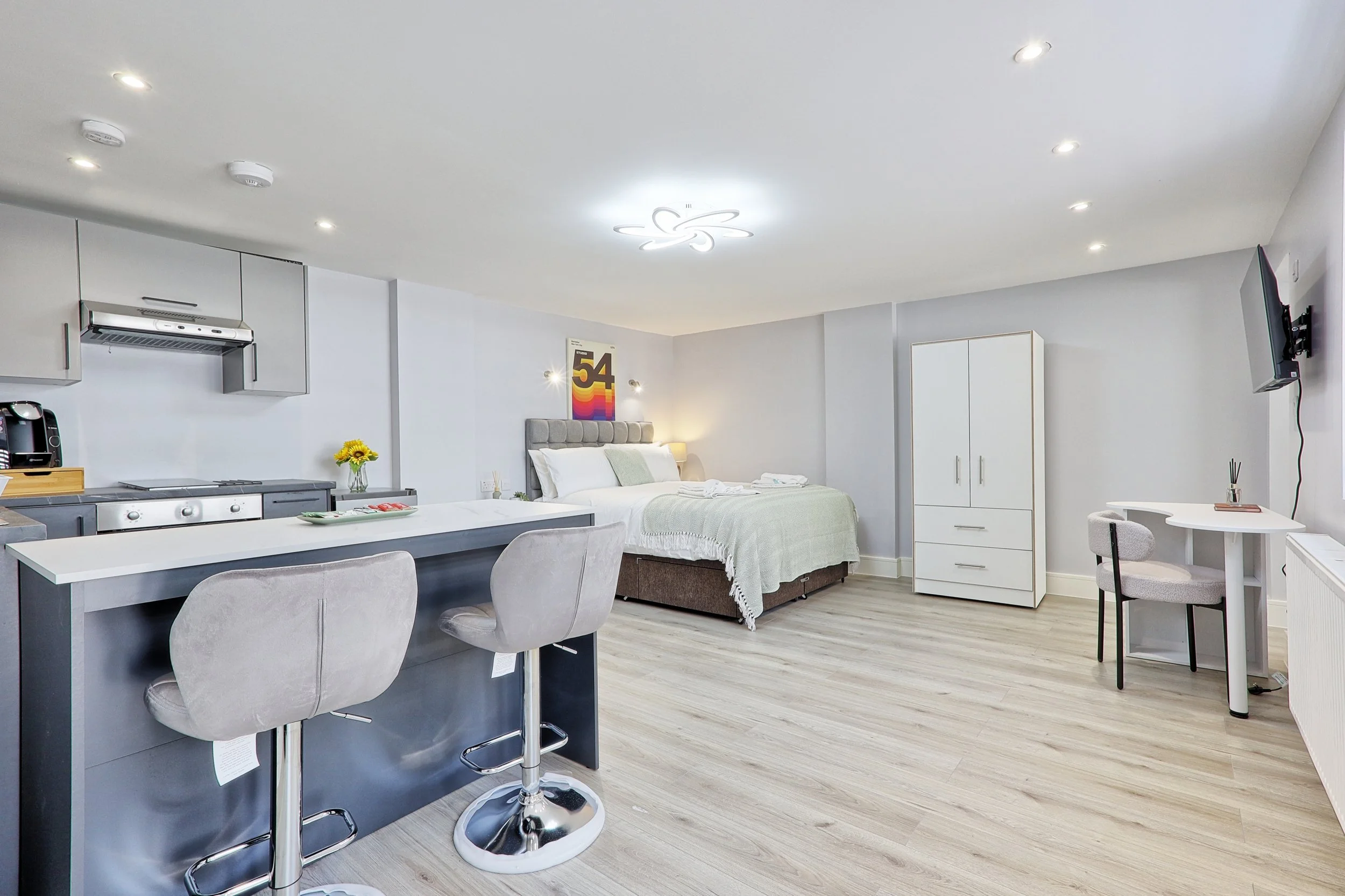 Modern studio apartment with kitchen area, small table, bed, wardrobe, and wall-mounted TV.